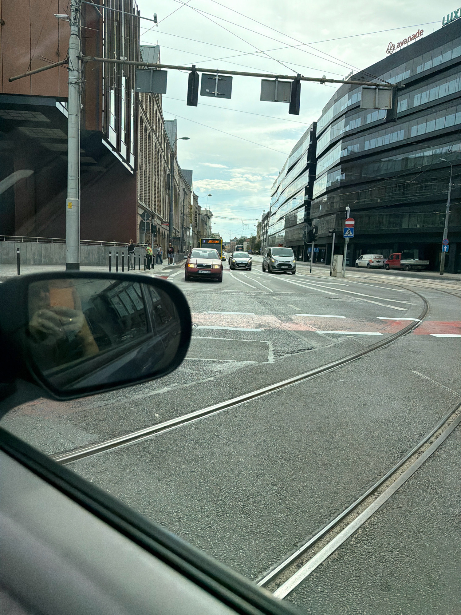 The image depicts a street scene in Wrocław, Poland, with a focus on tram tracks and the surrounding urban environment. The perspective is from inside a vehicle, looking forward and to the right. In the foreground, the road surface is visible, with prominent tram tracks running through it. The tracks are parallel and appear to be embedded in asphalt.
To the right, a modern, multi-story building with a curved, glass facade dominates the skyline. The building features reflective windows that mirror the sky. A sign reading "avanade" is visible near the top of this building.
To the left, another building with a brick facade and arched windows lines the street. A traffic light pole with traffic signals is present on the left side of the road. Several pedestrians are visible standing on the sidewalk in front of the brick building.
In the middle ground, several vehicles are present. A red Skoda passenger car is in the foreground, facing away from the viewer. Further down the street, a bus with the text "N LITEWSKA" is visible, as well as other cars and a white van.
A side mirror of the vehicle from which the photo is taken is visible in the lower left portion of the frame, reflecting a person holding a phone. The sky is overcast with clouds. The overall impression is a typical urban street with active traffic and pedestrian activity.