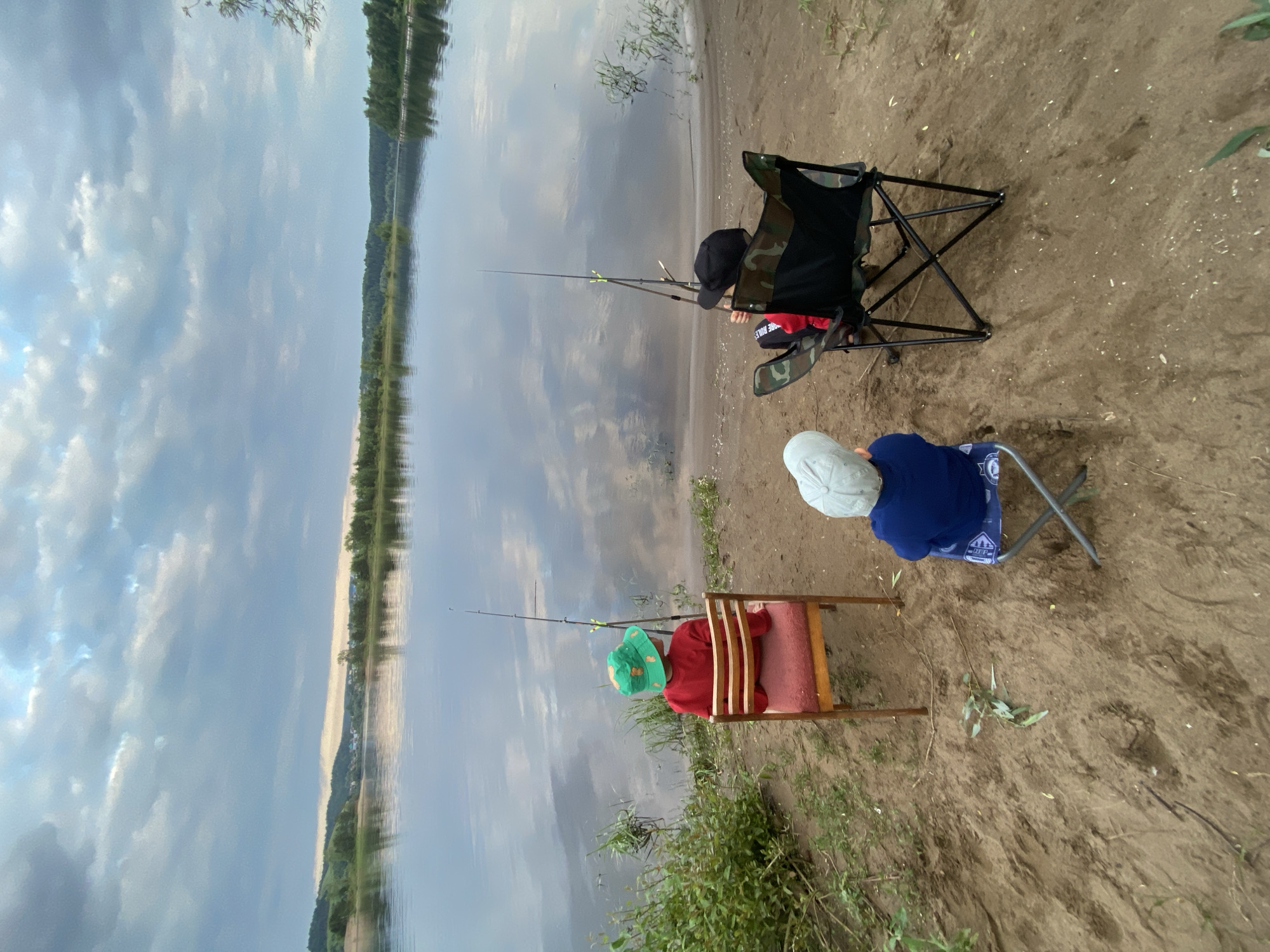Two children are fishing on the bank of a wide, calm river. The child in the foreground wears a red shirt and a green, patterned sun hat. They are sitting in a wooden chair with a red cushion. The second child, further back, wears a blue shirt and a light-colored sun hat, and is seated in a camouflaged camping chair. Both children have fishing rods in the water. The river reflects the cloudy sky, which is a mix of blues and grays. The far bank of the river is lined with trees and appears to have a light-colored sandy beach. The ground where the children are sitting is sandy with patches of green grass and vegetation. The overall scene suggests a peaceful, outdoor activity. The time of day appears to be late afternoon or early evening, given the soft lighting and cloud cover.