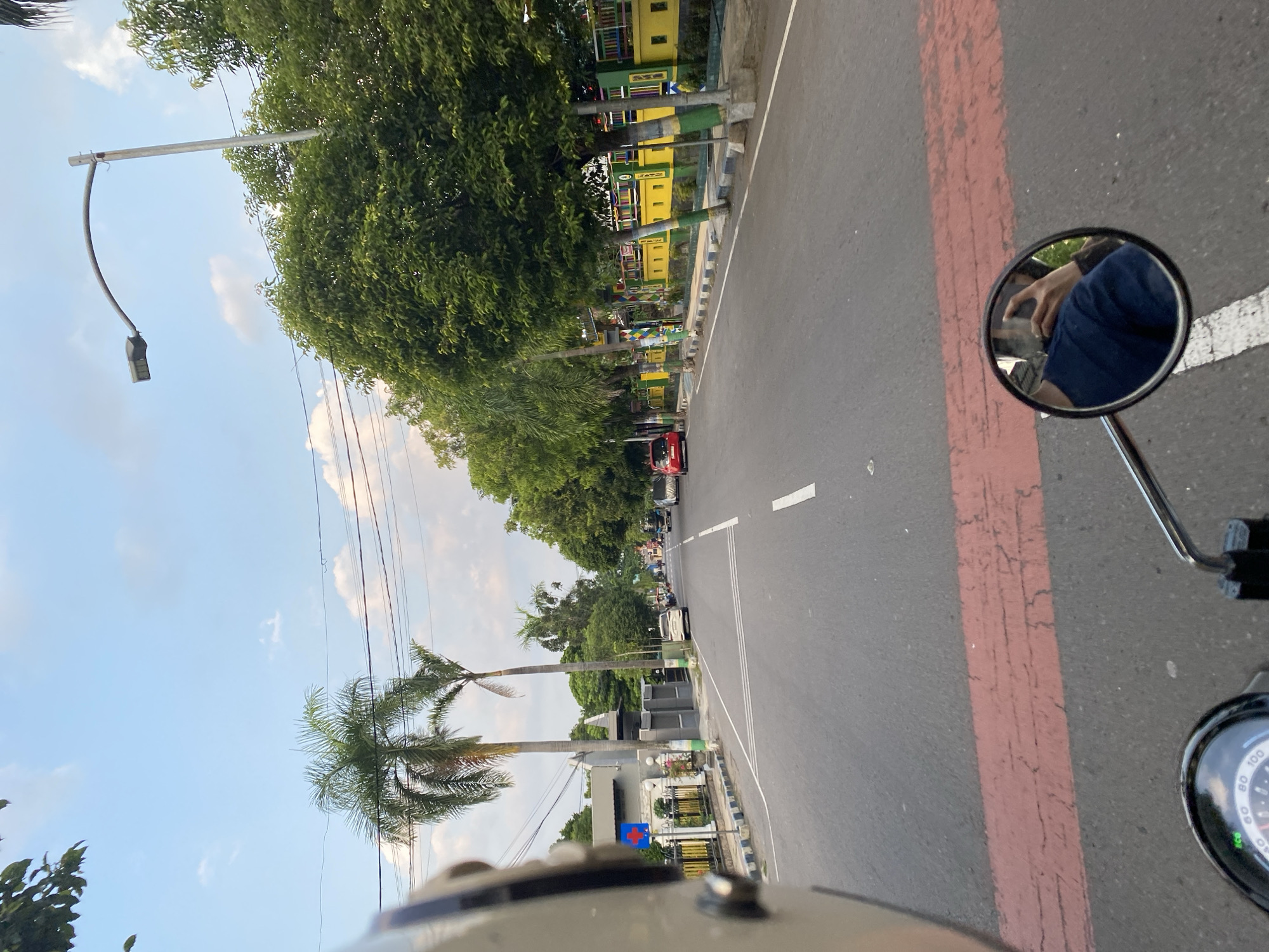 The image presents a first-person perspective from a motorcycle rider traveling along a paved road in Madiun, Indonesia. The scene depicts a clear, bright day, likely late afternoon, with a blue sky dotted with scattered white clouds.
The asphalt road features distinct markings, including a red stripe parallel to a white line on the right side, and a solid white line indicating traffic separation. The road is flanked by abundant greenery, consisting of lush trees and several tall palm trees, suggesting a tropical or subtropical environment. On the right side of the road, beyond the trees, colorful buildings or barriers painted in vibrant yellow and green are visible. Further down the street, other vehicles, including a red car, can be seen. A street lamp with a distinctive curved arm extends over the left side of the road.
In the immediate foreground, the top edge of a helmet or part of the motorcycle's frame is visible at the bottom of the image. A circular rearview mirror on the right prominently reflects the rider's hand and arm, dressed in blue, confirming the vantage point. A portion of the motorcycle's instrument cluster or speedometer is partially visible just below the mirror. Utility poles and power lines are visible against the sky on the left. Further down the road on the left, a blue sign with a white cross, typically denoting a medical facility or first aid, is discernible. The scene conveys a sense of a pleasant journey through a lively urban or suburban setting.