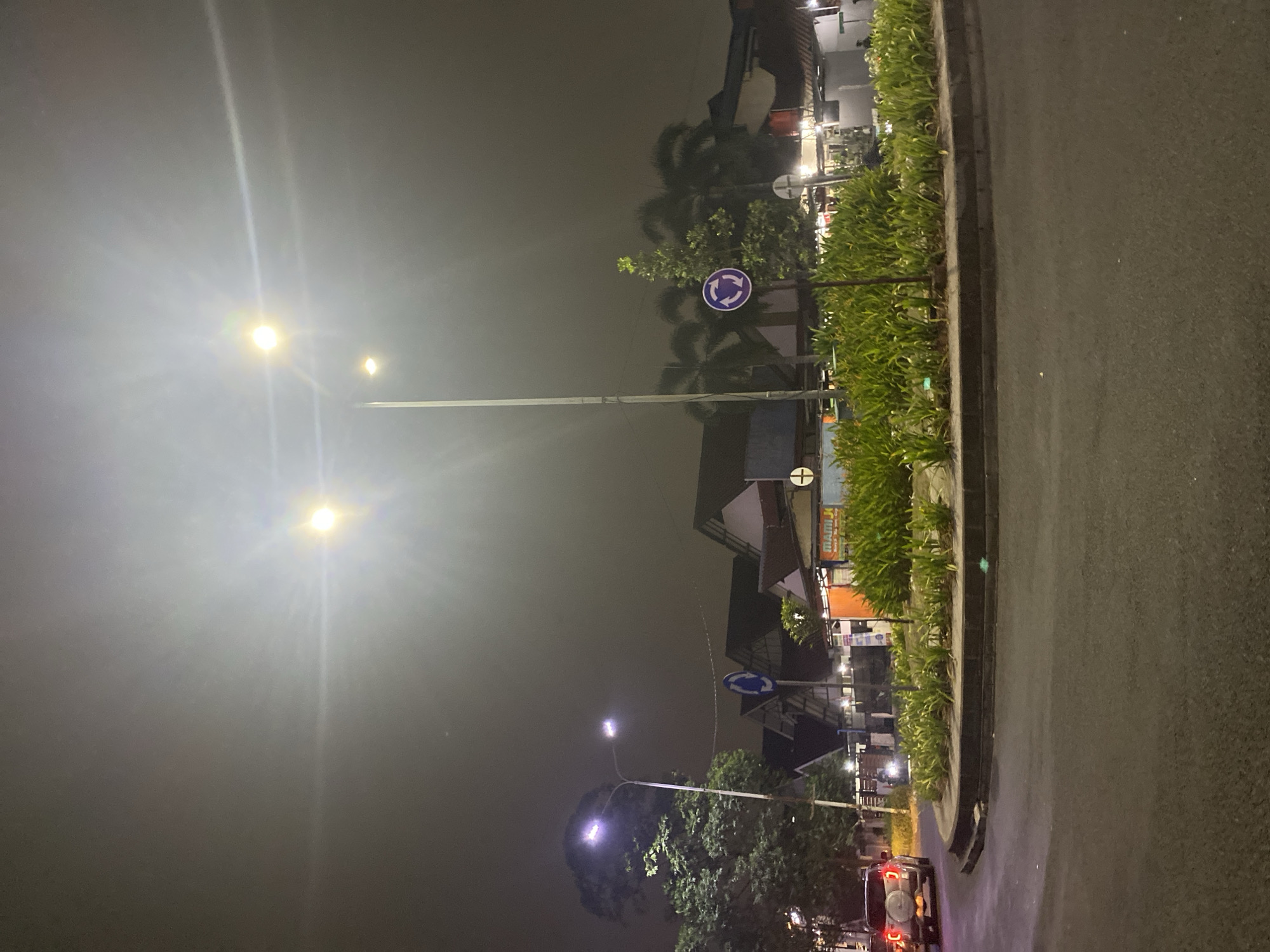 The image depicts a roundabout at night in Tangerang, Indonesia. Streetlights illuminate the scene, casting a glow on the wet asphalt road and a central garden bed filled with lush green plants. Traffic signs indicating a roundabout are visible on poles. In the background, several buildings with pitched roofs are illuminated. A silver SUV is seen entering the roundabout, and a motorcycle is also present. The sky is dark and hazy, suggesting a humid or polluted atmosphere. The overall impression is one of a typical urban nighttime environment in a tropical region.
