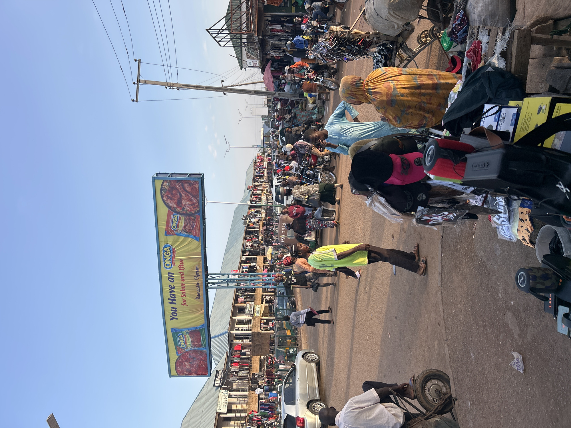 The image shows a bustling street scene in Jos, Nigeria, characterized by a crowded marketplace. A large billboard advertises ONGA seasoning with the text "You Have an ONGA for Sahur and Iftar Ramadan Karëëm." Numerous people are visible, many on motorcycles, amidst market stalls selling various goods. A few pedestrians walk along the road, while others are engaged in commerce near the stalls. A silver car is parked by the roadside. Street vendors are selling wares next to the photographer. The image is taken on a sunny day with a clear, blue sky.