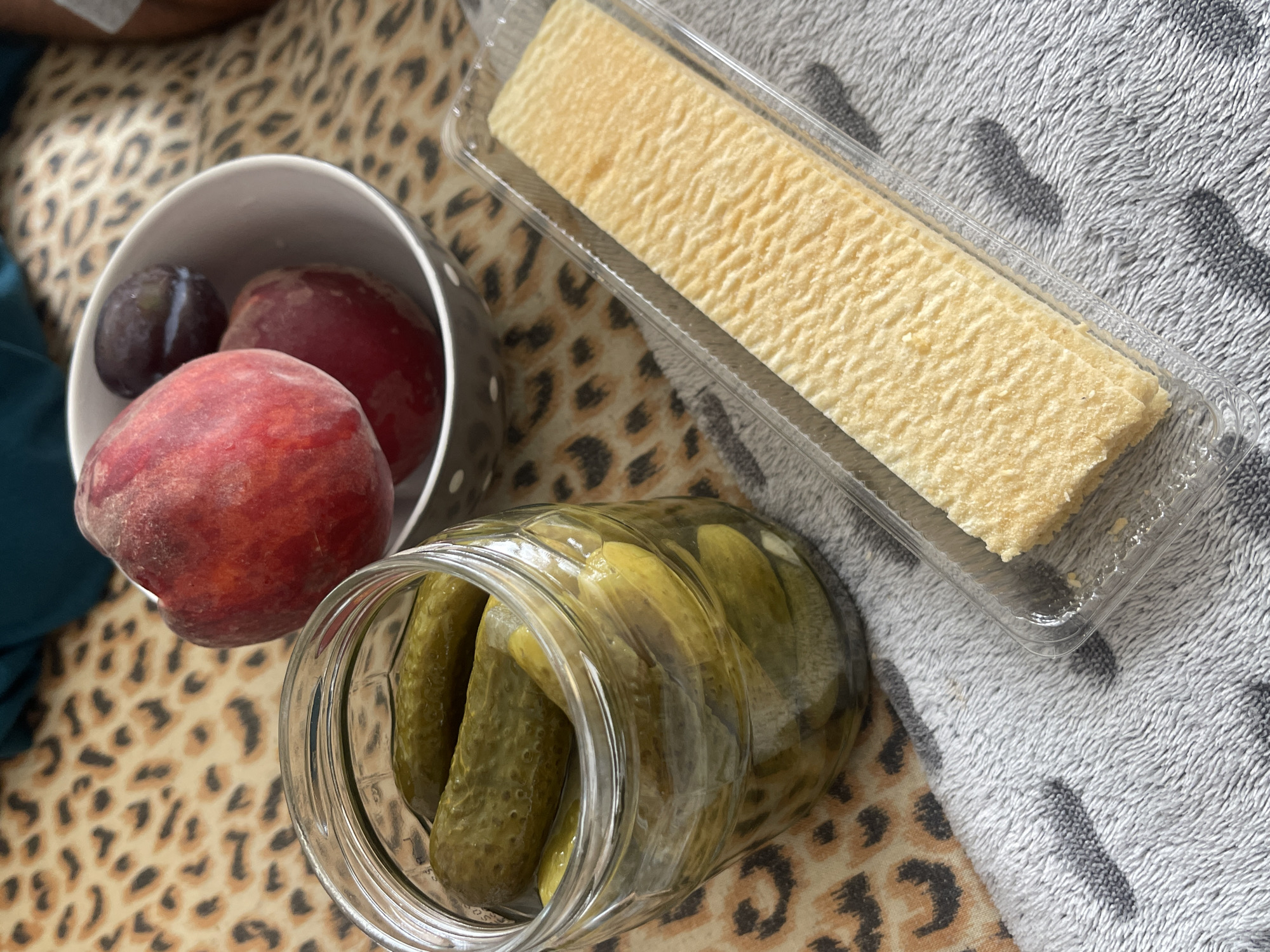 A close-up, overhead shot shows a selection of food items arranged on a leopard-print fabric and a grey textured cloth. In a clear glass jar, several green pickles are visible. To the right of the jar, a grey and white polka-dotted bowl contains two red peaches and one dark plum. In the foreground, a rectangular wafer is presented in a clear plastic container. The scene is lit by natural light. This image does not contain information about the city of Varna, Bulgaria.