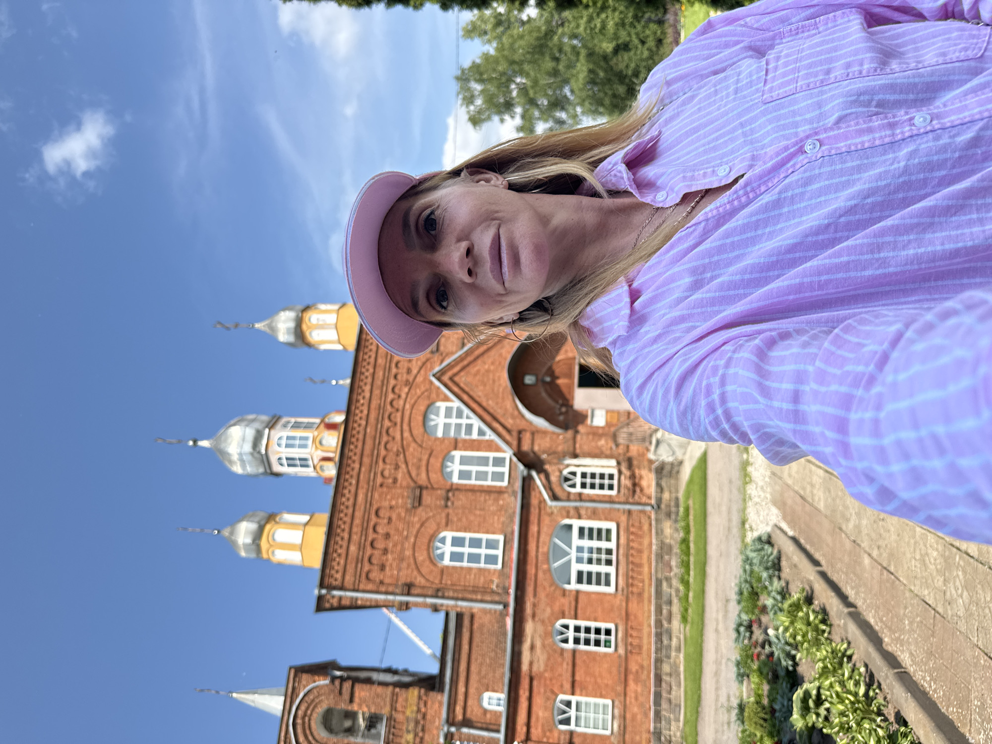 A woman wearing a pink striped shirt and a pink baseball cap takes a selfie in front of a red brick church with silver domes. The church has arched windows and appears to be an Orthodox Christian church. There are green trees and a blue sky in the background. The woman is smiling and looking at the camera. The church is identified as being located in Chudovo, Russia.