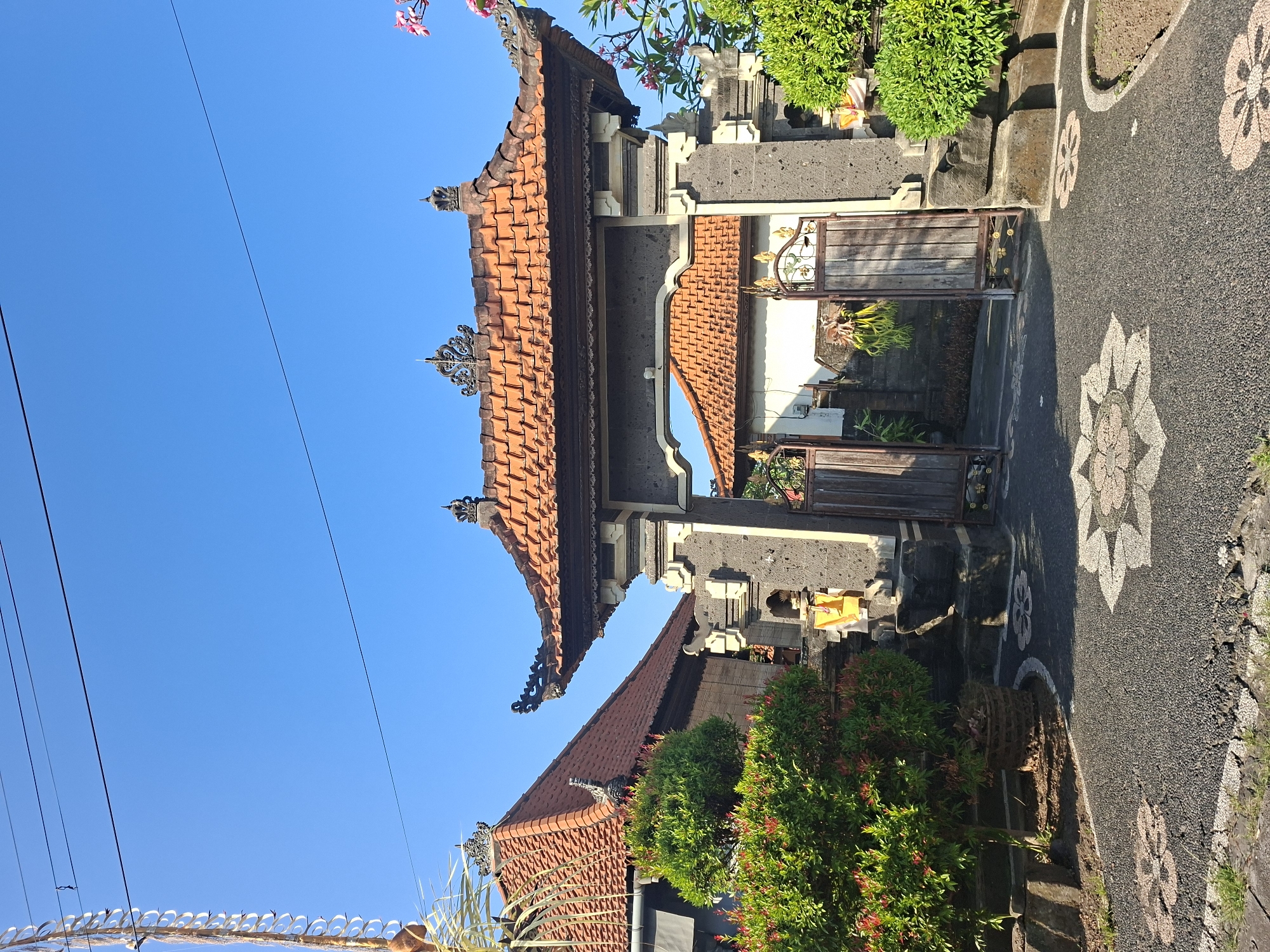The image shows an ornate stone gateway with a tiled roof, likely part of a traditional Balinese compound, in the village of Blahbatuh, Indonesia. The gateway is constructed from dark, rough-hewn stone blocks, topped with a dark wooden frame and an orange-tiled, multi-tiered roof. Decorative carvings adorn the top of the gateway structure, and two weathered wooden gates, one partially open, stand within the archway.
To the left of the gateway, a lush green tree with red-tinged leaves is planted in a woven basket. Behind the tree, a building with a similar orange-tiled roof is visible, its exterior walls appearing to be a lighter color. The pathway leading up to the gate is paved with dark gravel and features inlaid patterns of light-colored stone creating floral motifs.
To the right of the gateway, another well-maintained green shrubbery is present, partially obscuring the stone structure. In the background, beyond the gateway, another building with an orange-tiled roof is visible. The scene is bathed in bright sunlight, casting clear shadows, and the sky above is a vibrant, cloudless blue. There are no people or animals visible in the image. The overall impression is one of a well-kept, culturally rich entrance to a residential or religious compound.