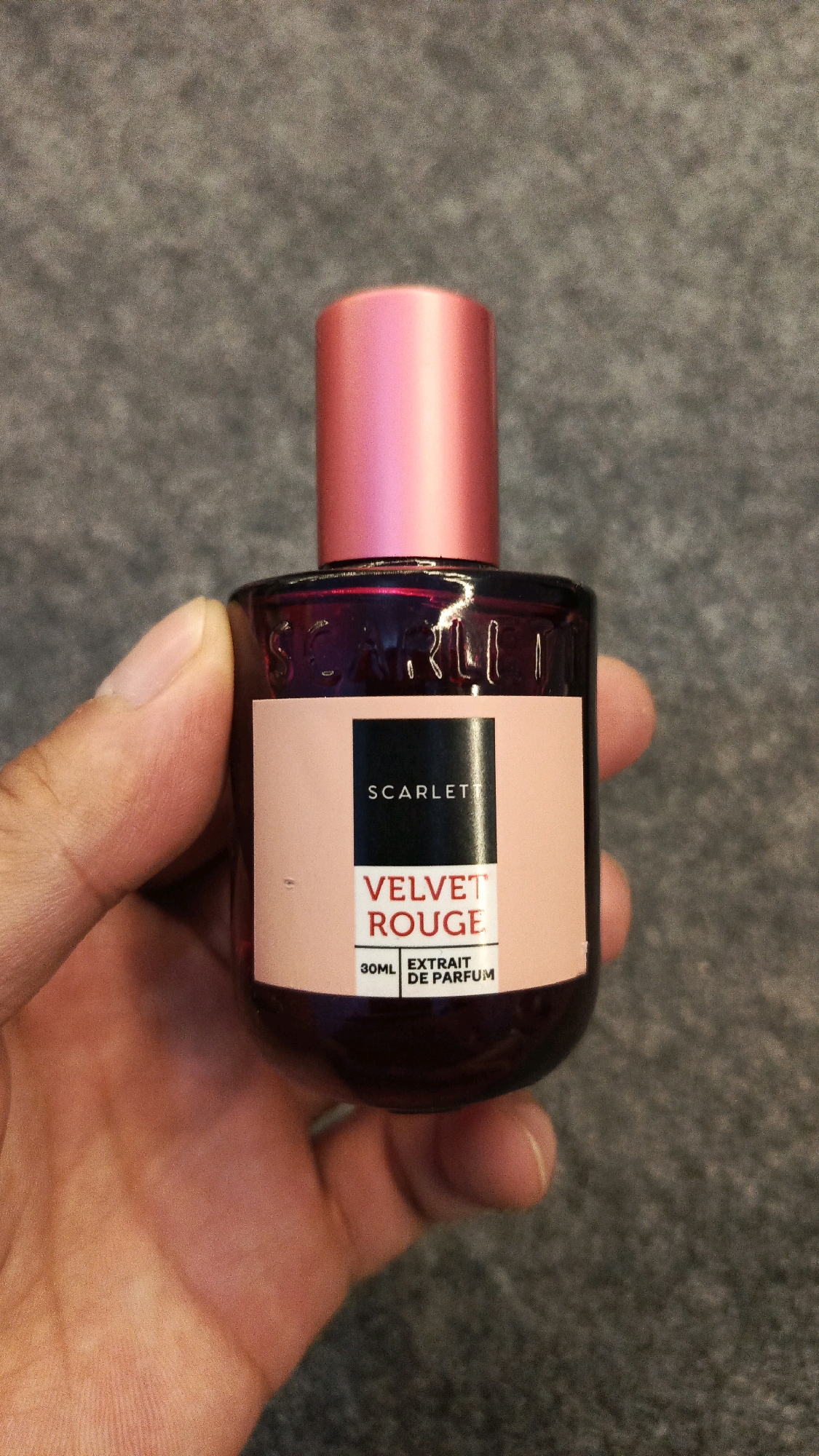 A person's hand is holding a small, dark glass perfume bottle with a pink cap. The bottle has a peach-colored label with black text that reads "SCARLET" at the top, followed by "VELVET ROUGE" in larger red text. Below this, in smaller text, are "30ML" and "EXTRAIT DE PARFUM". The background is a grey, textured surface.