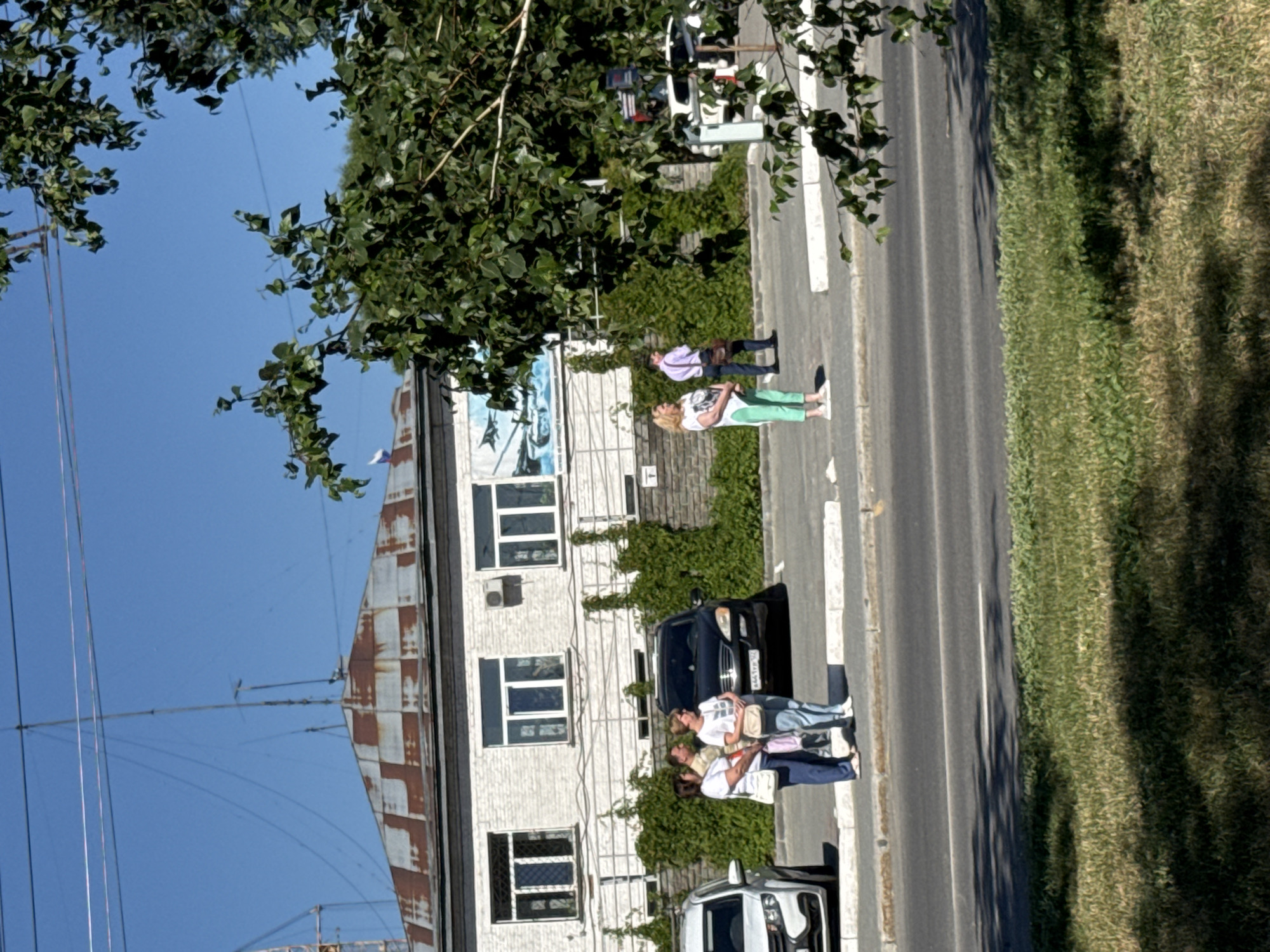 The image shows a bright, sunny day in Barnaul, Russia, with a clear blue sky. In the foreground, a grassy area with dappled sunlight is visible. Behind it, a paved road with a white line marking is present. A sidewalk runs alongside the road, separated by a low concrete barrier.
On the sidewalk, several people are standing near parked cars. To the left, two women in white shirts and dark pants are conversing, with a white car parked next to them. A black SUV is parked to their right. Further down the sidewalk, a woman in a white shirt and green pants stands with a man in a purple shirt and dark pants, carrying a briefcase.
Behind the sidewalk and people, a white building with a corrugated metal roof is visible. The building has several windows and is partially covered by ivy. A mural depicting a tank and an aircraft is on the building's facade. Electrical wires are strung across the top of the image, and a tall antenna or radio tower is situated above the building. A small Russian flag is flying from the roof of the building. The scene appears to be a public street or square in the city.