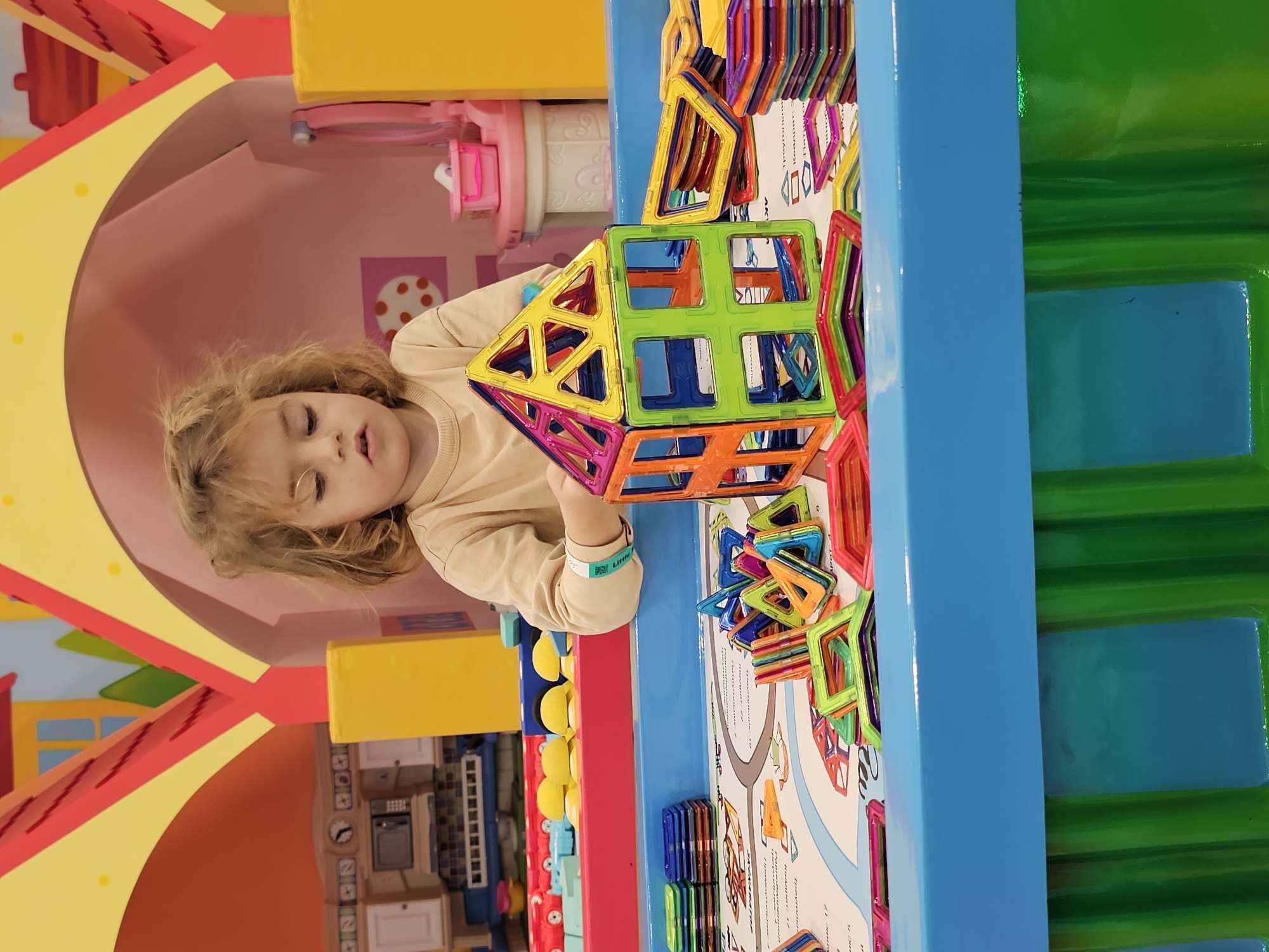 A young child with light blond hair, wearing a light beige long-sleeved shirt and a blue wristband reading "Little," is holding a multicolored geometric structure of magnetic tiles, seemingly inside an indoor playspace in the city of Khimki, Russia. The structure consists of colorful squares and triangles in red, orange, yellow, green, and blue. The child is positioned by a play table with a blue edge filled with various magnetic tiles. A white play mat with drawings lies on the table. To the left of the child, there's a play kitchen set against a pink-walled backdrop with polka dots, under a yellow and red play structure. Further to the right is a green and blue structure. The overall lighting suggests an indoor environment.
