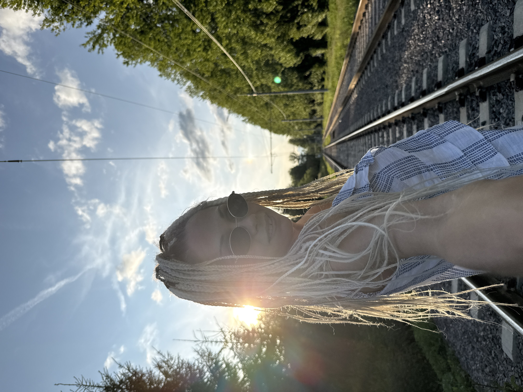 A blonde woman with numerous braids and sunglasses stands on a railway track, looking towards the camera. She is wearing a blue and white checkered top. The sun is setting behind her, casting a golden light on her hair. The railway tracks stretch into the distance, lined with trees on either side. Overhead, power lines are visible against a blue sky with scattered clouds. The scene appears to be located in Rusanovo, Russia.