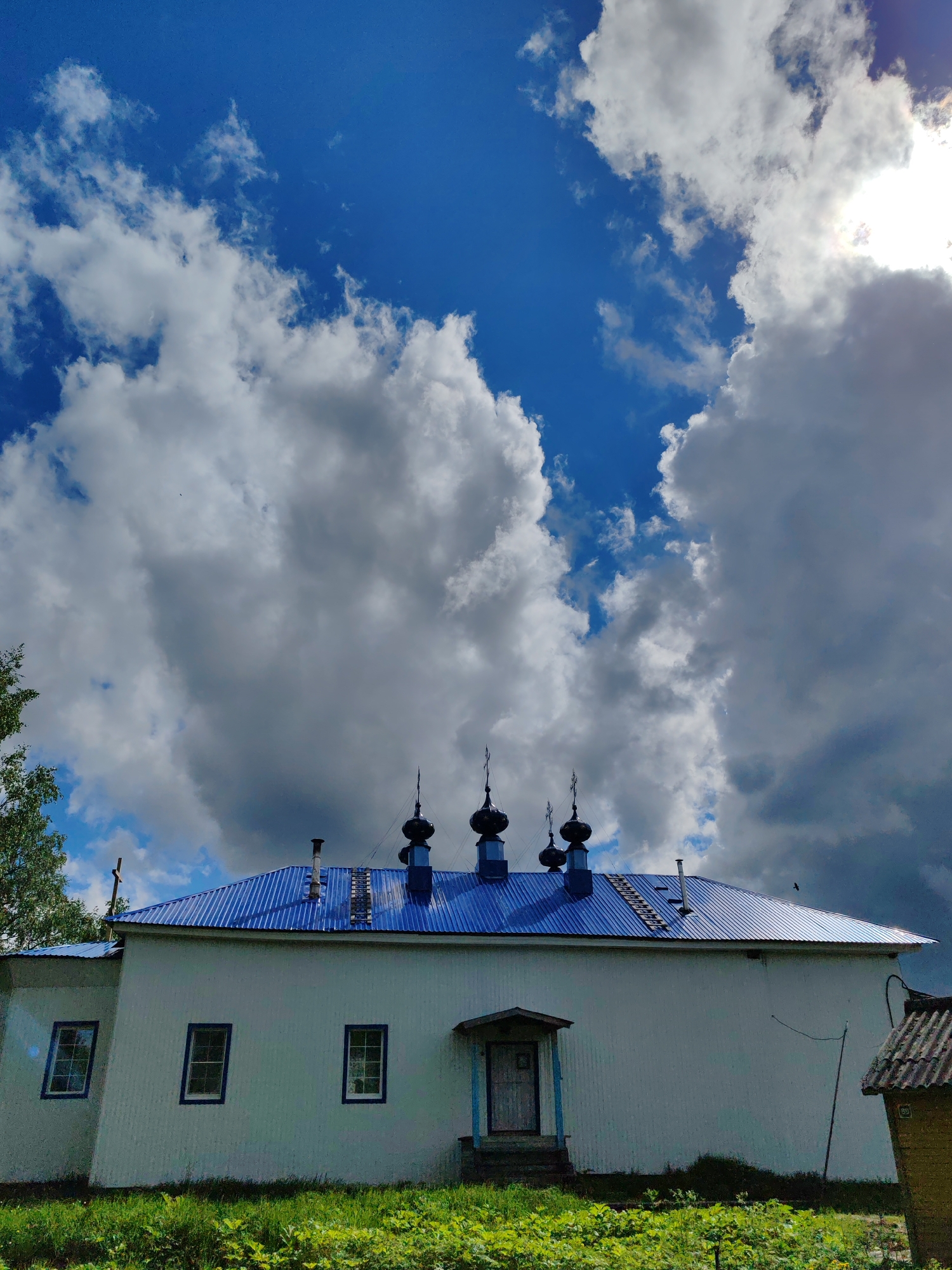 The image shows a white, single-story building with a blue metal roof and four dark onion domes atop it. Three windows are visible on the side of the building, each with blue trim. A small porch with a door is in the center. The roof has a chimney and what appears to be a ladder-like structure. Green grass and vegetation are in the foreground. The sky is blue with large, puffy white and gray clouds. The location is Чухлэм, Russia.
