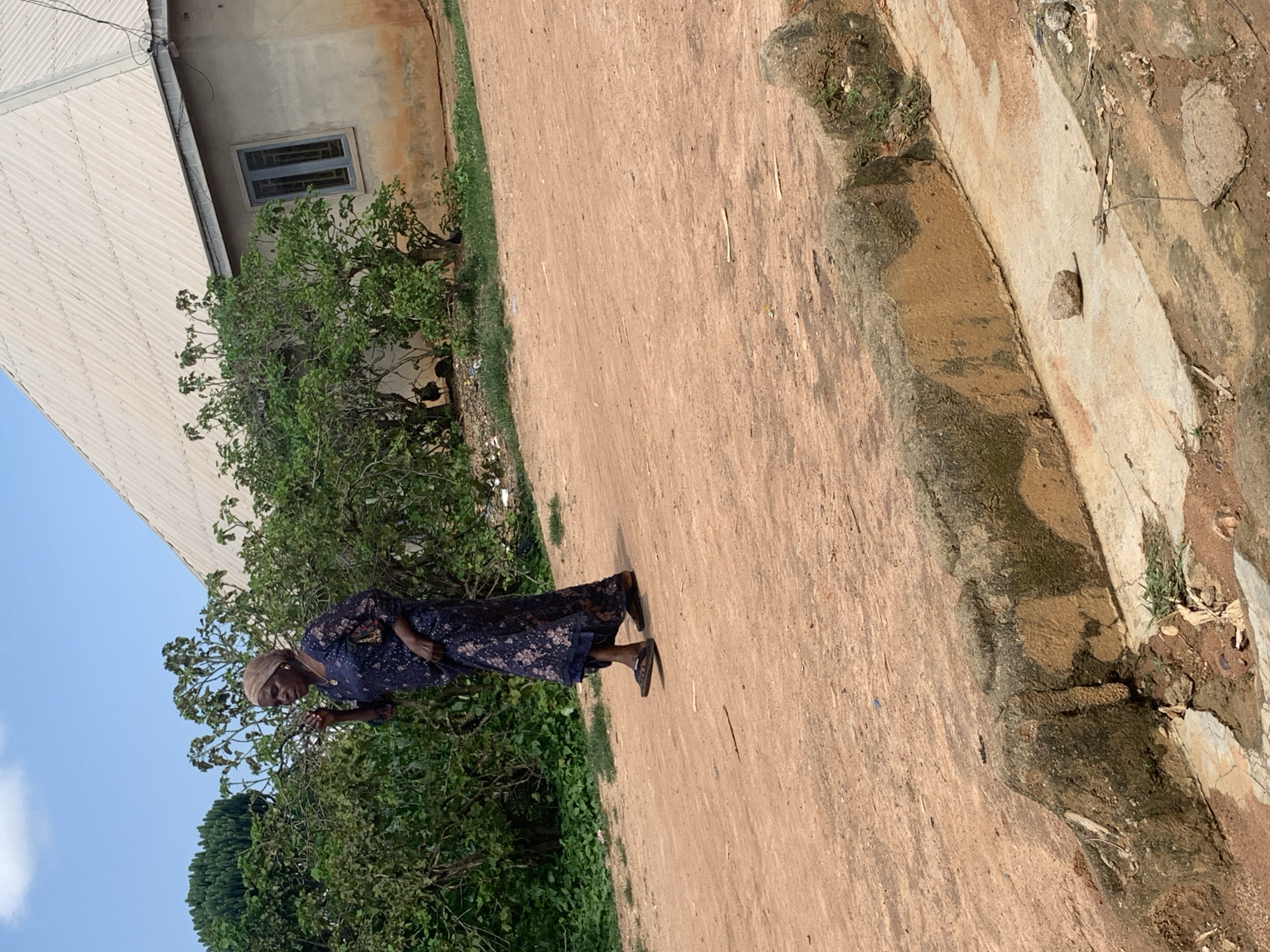 A middle-aged Black woman wearing a dark blue floral dress and a light brown headscarf is walking on a dirt road. The woman is facing to the left of the frame and her right hand is raised to her mouth. She is wearing dark sandals. Behind the woman, there is a building with a light-colored corrugated metal roof and a grey-framed window. Green bushes and trees are visible around the building. The road is made of reddish-brown dirt. A low, rough concrete structure with moss and dirt is in the foreground. The sky is blue with some white clouds.