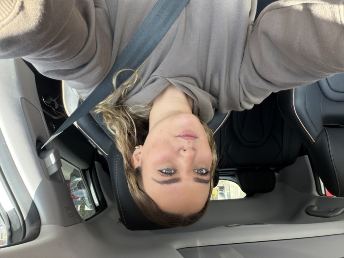 A young woman is captured in a close-up selfie inside a car, looking directly at the camera with a neutral expression. She has light-colored, possibly blonde or light brown, wavy hair that frames her face. She is wearing a light brown or grey hooded sweatshirt and has a gold-colored hoop earring visible in her right ear. A dark seatbelt crosses her chest and shoulder, with the word "AIRBAG" faintly visible on what appears to be part of the seat or pillar near her left shoulder.
The setting is the interior of a car, likely the front passenger seat, during daylight hours, indicated by the bright, clear lighting. The car's upholstery is dark, possibly black leather or fabric. Through the car windows, reflections of other vehicles and an urban or suburban outdoor environment are visible, suggesting the car is either parked or stationary. The image implies a sunny day. Given the location context, this scene is taking place in Puebla City, Mexico.