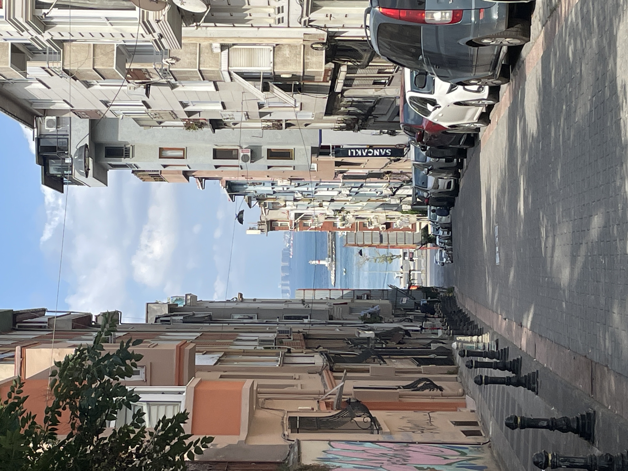 The image captures a street scene in Kadıköy, Turkey, looking down a narrow road lined with multi-story buildings. The architecture features a mix of styles, with balconies, awnings, and air conditioning units visible on the facades. The buildings have various colors, including peach, grey, and white. The street is paved with cobblestones and lined with black bollards on the left side. Several cars are parked along the right side of the street, including a dark grey van in the foreground, a white sedan, and other sedans and SUVs. In the distance, the street opens up to a view of the sea and a lighthouse. The sky is blue with scattered clouds. Graffiti is visible on the wall of a building on the left side of the street. A banner with Turkish text, likely advertising a business, hangs vertically from one of the buildings. The scene appears to be a residential or mixed-use area in a coastal city.