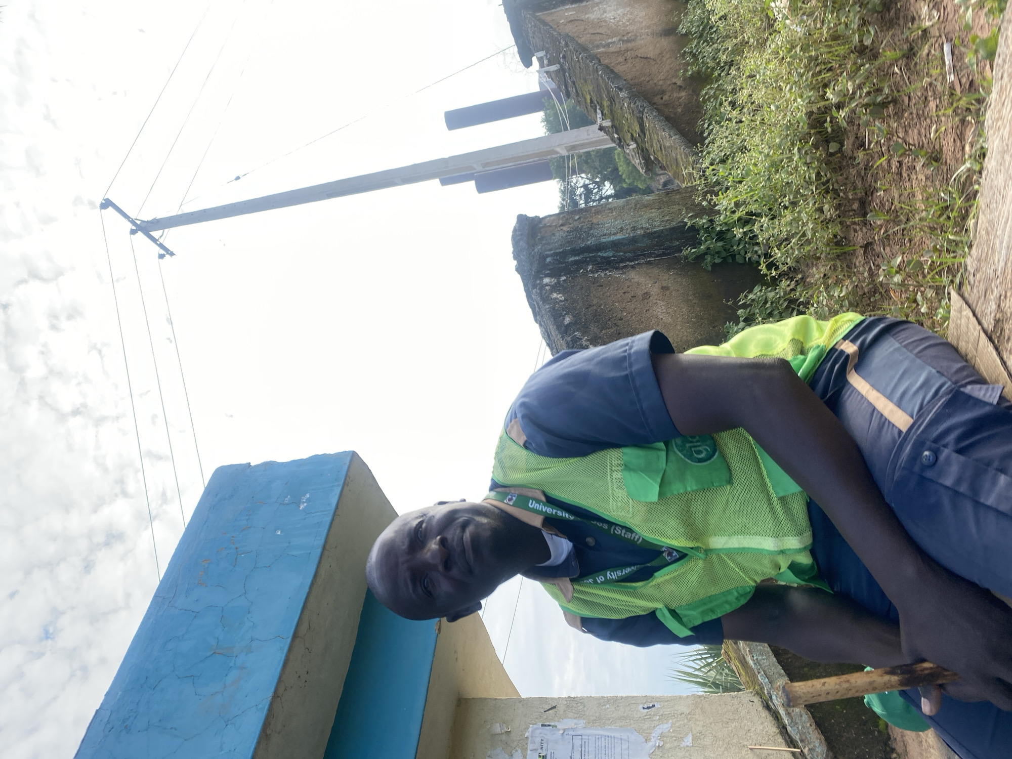 An outdoor, medium shot shows a Black man sitting outdoors and holding a wooden stick. He is wearing a dark blue shirt, a bright green safety vest with the brand name "Glo" visible, and dark blue pants with a tan stripe. He has a lanyard with a green and white tag that reads "University of Jos (Staff)". He is sitting on a wooden surface, and behind him, to the left, is a partially visible blue building with cracks in its paint. To the right, there is a concrete structure with moss growing on it, and in the background, a wooden utility pole with wires runs vertically. The environment appears to be somewhat overgrown with vegetation. The man is looking slightly to his right. The location is identified as Rafin Par, Nigeria.