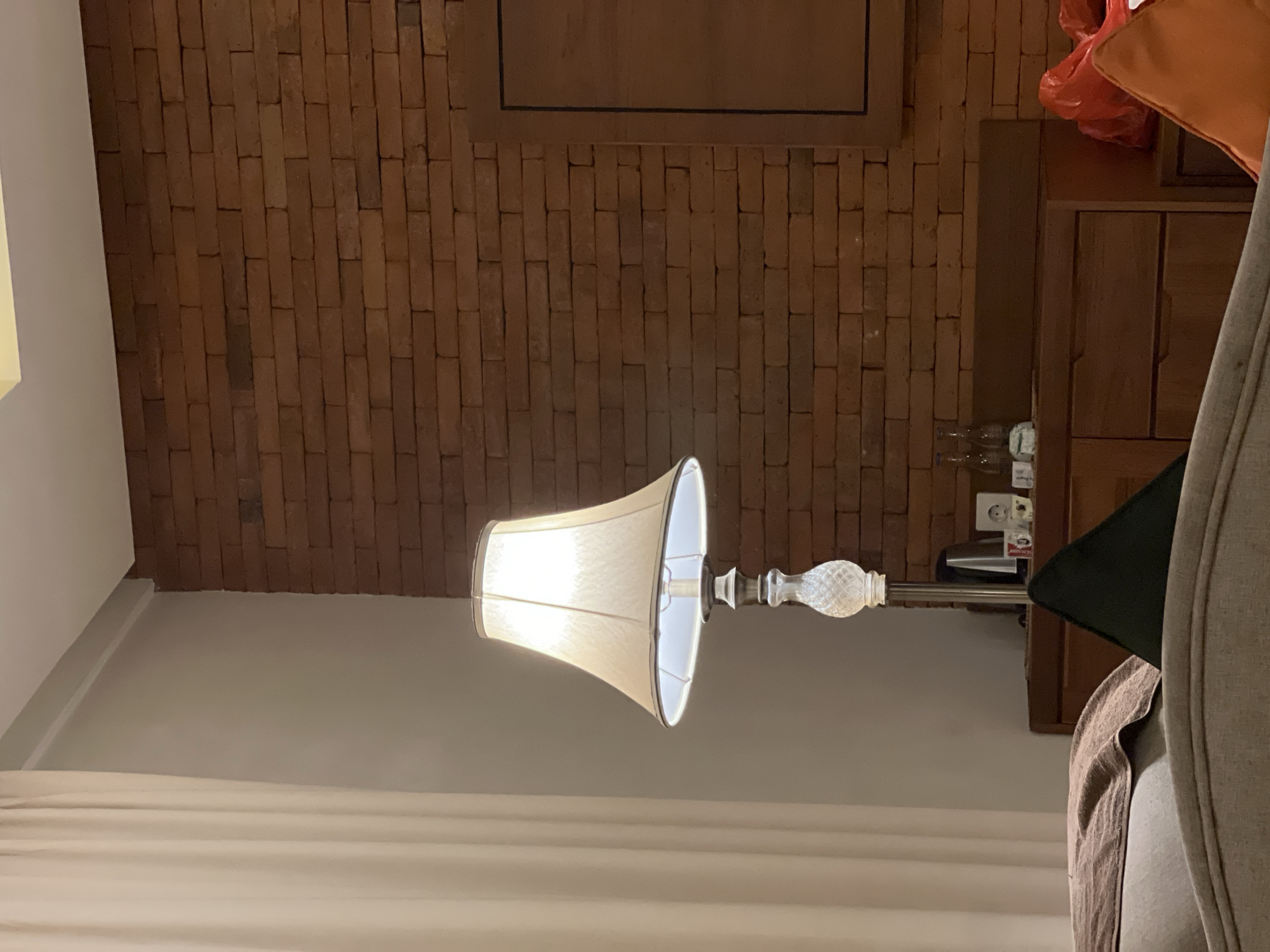 An interior scene, likely a hotel room or guesthouse in Ubud District, Indonesia, is captured under artificial light. The central focus is an upright floor lamp featuring a cream-colored fabric shade that emits a warm glow, suggesting the photo was taken in the evening or at night. The lamp has a decorative, possibly crystal or carved glass, finial on its stem.
Behind the lamp, a prominent wall of exposed red brick provides a rustic backdrop. Above the brick wall, a dark wooden panel or frame is partially visible. To the right of the frame, a dark wooden cabinet or bedside table stands, upon which a crumpled red plastic bag is placed. Below the cabinet, two clear plastic water bottles are neatly stacked. A white power outlet, consistent with types commonly found in Indonesia, is visible on the brick wall to the right.
The foreground and left side of the image show a light-colored wall meeting a white, possibly sheer, curtain or bedspread. Part of a soft furnishing, likely an armchair or headboard with a light brown throw, is partially visible on the far right. The room appears unoccupied, with no specific activity taking place, conveying a sense of quiet and stillness. There is no visible text.