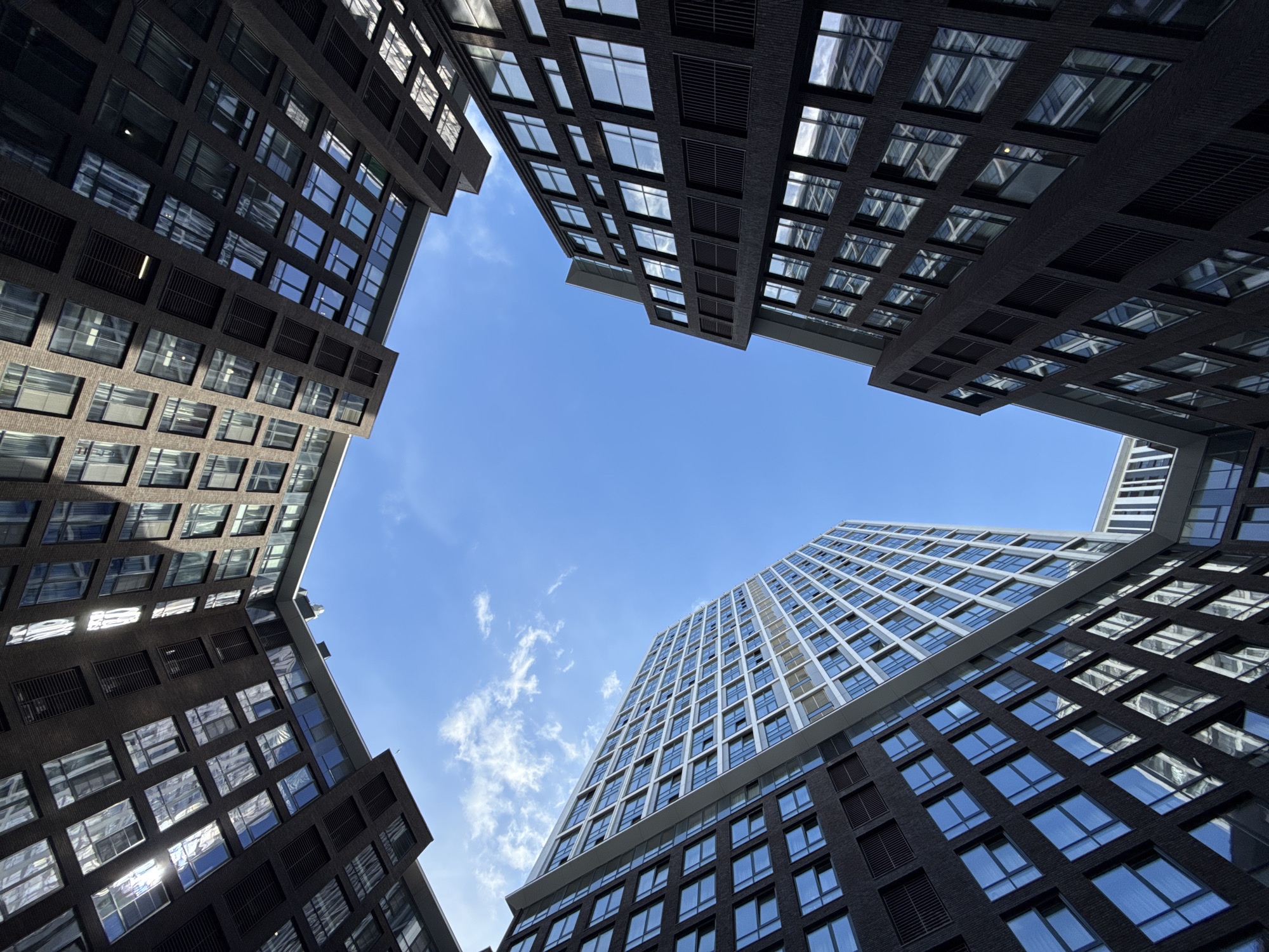 From a ground-level perspective, the image captures a dramatic view of four tall, modern buildings encircling a square of blue sky with scattered clouds. The buildings are constructed with dark brick or stone facades and feature numerous rectangular windows, many reflecting the sky. The architectural style is contemporary, with clean lines and a grid-like pattern of windows. The buildings are positioned at oblique angles, converging toward the center of the frame, creating a sense of depth and enclosure. The location is identified as Moscow, Russia based on the distinctive modern architecture. There are no people visible in the image.