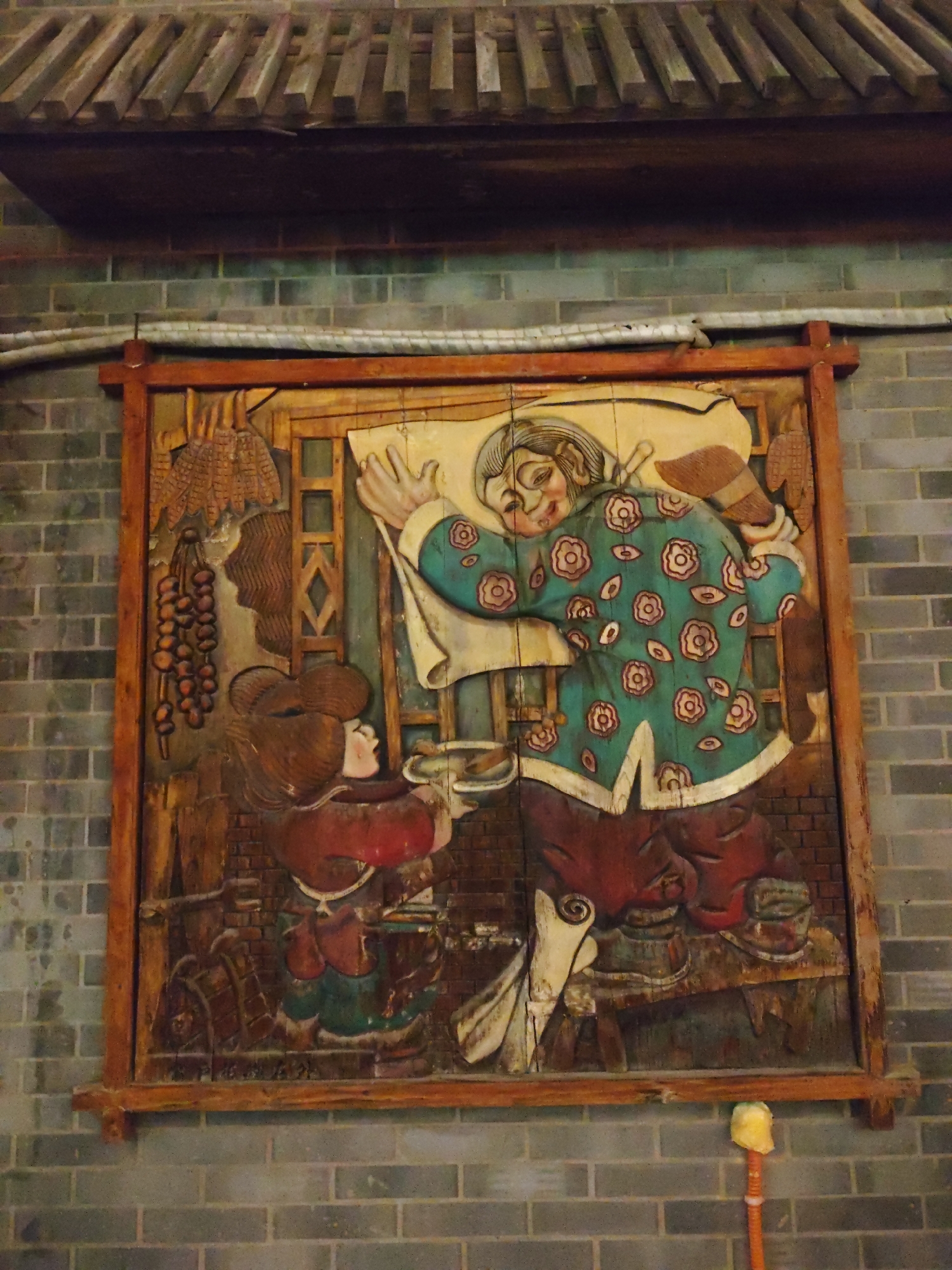 A carved wooden relief depicting a scene with two individuals, one larger and one smaller, is mounted on a brick wall in Lindian, China. The larger figure, centrally located, wears a turquoise robe adorned with pink and white floral patterns and dark red trousers. They are depicted with a smiling expression and appear to be carrying or moving a large, flat object over their shoulder. A smaller figure, positioned to the left of the larger figure, wears a red top and dark trousers, and is facing the larger figure, appearing to be engaged in an activity with them. To the left of the figures, a cluster of round, orange objects hangs, possibly representing fruit or vegetables. The carving is framed by a wooden border. The background of the carving features textures suggesting wooden planks and architectural elements. Above the carving, a slatted wooden structure is visible. The brick wall itself is constructed of gray-blue bricks.