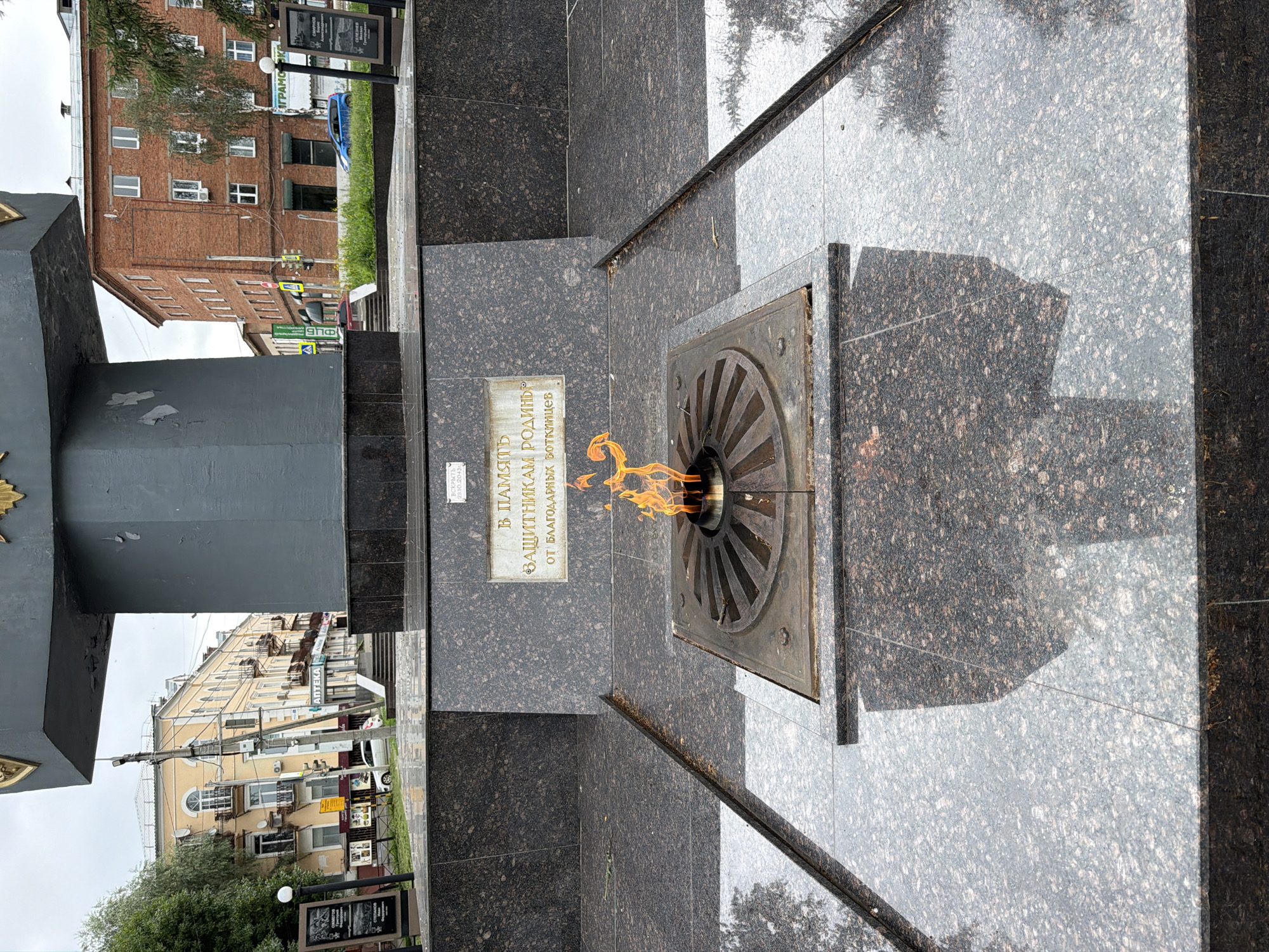 The image shows a memorial with an eternal flame in the city of Votkinsk, Russia. The memorial is made of dark granite and features a plaque with an inscription in Russian. The eternal flame is burning in a circular metal grate. In the background, there are buildings, trees, and a cloudy sky. The overall scene is solemn and reflective.