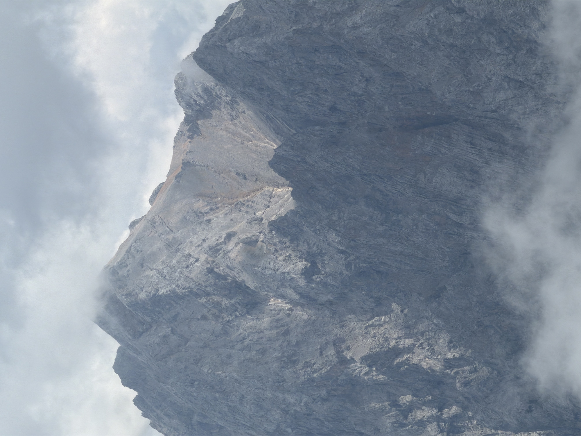 The image shows a close-up view of a rugged, gray mountain peak under a cloudy sky. The mountain's face is characterized by steep slopes, sharp ridges, and a rough, rocky texture. Patches of light and shadow highlight the contours and crevices of the terrain. Wisps of fog or clouds are present at the lower portions of the mountain, partially obscuring the base. The overall scene conveys a sense of natural grandeur and remoteness. This mountain is likely part of the Bavarian Alps, a region that includes the municipality of Garmisch-Partenkirchen, Germany. No people or specific objects are discernible within the frame of the mountain itself.