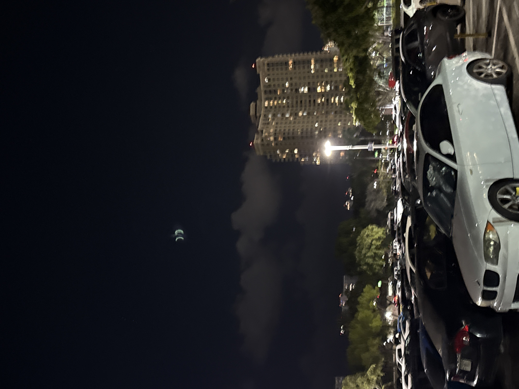 The image depicts a nighttime scene in what appears to be a parking lot in Aventura, Florida, United States. A tall building with numerous illuminated windows is visible in the background, suggesting an urban environment. Several parked cars are arranged linearly, with a white sedan prominently positioned in the foreground. The sky is dark, with the presence of clouds and a faint celestial object. Trees and foliage are located along the edge of the parking area, indicating a partial park-like environment. The parking lot itself is well-lit with streetlights.