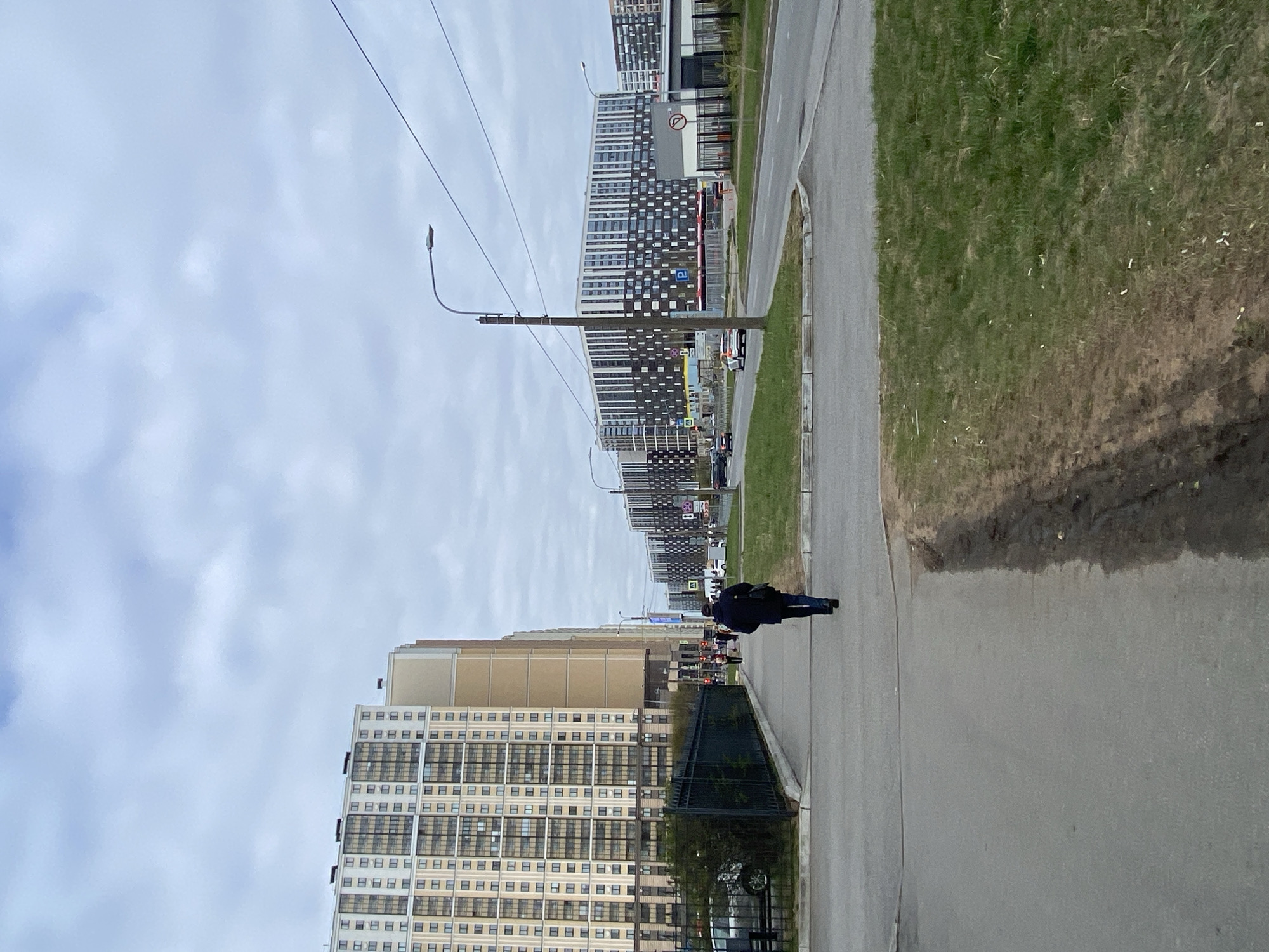 An outdoor image depicts a modern urban street scene in Saint Petersburg, Russia, under an overcast sky. The shot is taken from a slightly elevated perspective, looking down a long, straight road bordered by sidewalks and tall apartment buildings.
To the left, a prominent multi-story residential building dominates the foreground, featuring a light-colored facade with numerous rectangular windows, some dark and reflective. Further down the street on the left, other similar buildings extend into the distance.
A wide, paved sidewalk runs through the center of the frame, leading into the background. A person, dressed in a dark jacket and trousers, is walking away from the viewer on this sidewalk. To the right of the sidewalk is a paved road with a white painted line. Beyond the road, a strip of green grass with some scattered white debris separates it from another set of modern high-rise buildings. These buildings on the right display a more patterned facade, combining dark and light sections with many windows.
Several streetlights on tall poles are visible, stretching along the road, and power lines crisscross the cloudy sky. Near the buildings on the right, a circular traffic sign with a red border and a diagonal line (likely indicating "No Stopping" or "No Parking") and a blue parking sign with a white 'P' are discernible. The time appears to be daytime, likely a cloudy day, given the diffused light and lack of direct shadows. The overall scene is one of everyday activity in a newly developed residential area.