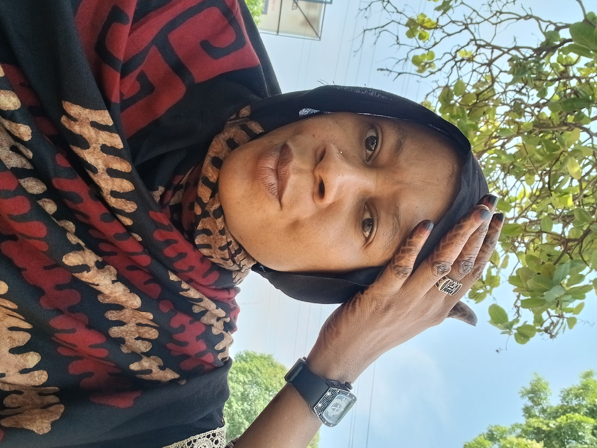 A close-up, angled portrait features a person, likely a woman, looking directly at the camera from an upward perspective. She has dark skin and a neutral expression, with dark eyes and a small nose piercing. Her right hand is raised to the side of her head, displaying faint mehndi (henna) patterns on her fingers and palm, along with a gold-colored ring with an openwork rectangular design on her ring finger.
She is wearing a head covering (hijab or scarf) that is primarily black, patterned with bold red and golden-tan abstract and geometric designs. A black watch with a distinctive rectangular, ornate silver-toned face and a black strap is visible on her left wrist. Part of her attire also shows a strip of delicate golden lace trim.
The setting is outdoors during the daytime, indicated by the bright, clear light blue sky and the vibrant green foliage of trees in the background. The low camera angle places the person against this natural backdrop. The overall mood is calm and contemplative. No discernible text is clearly visible on the watch face or ring. The scene is set in Shaba, Nigeria.