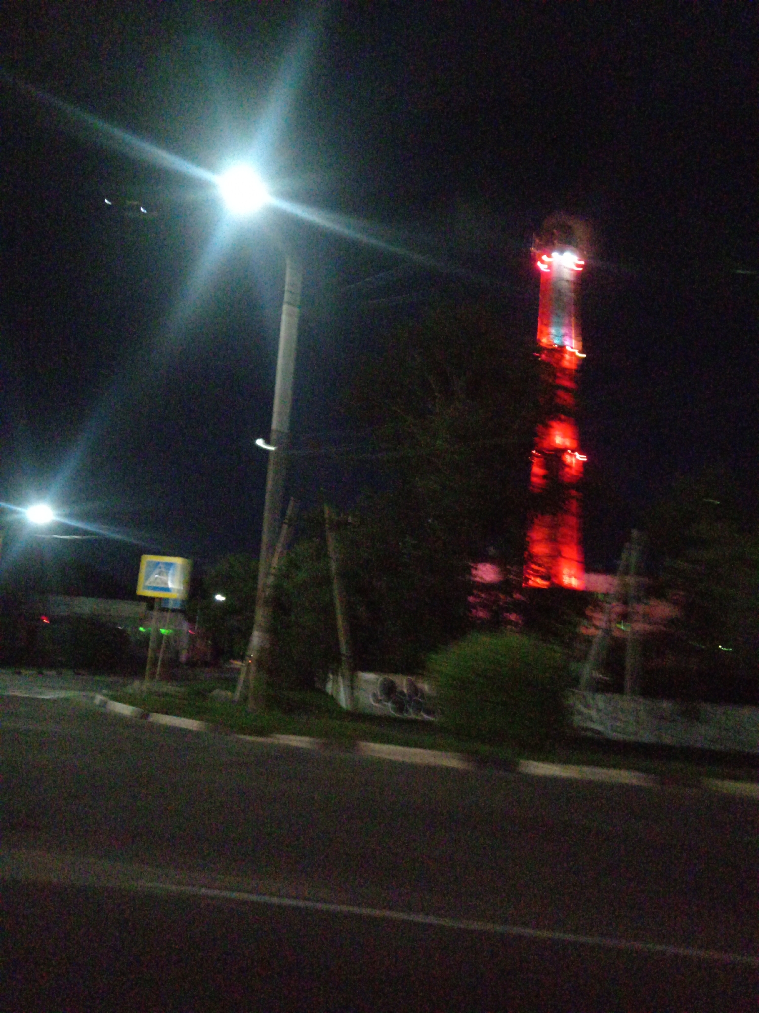 The image depicts a nighttime scene in the city of Belogorsk, Russia. A brightly lit street lamp dominates the left side of the frame, casting a strong glare. In the distance, a tall, illuminated structure, likely a radio tower or similar industrial facility, is visible, glowing with red and white lights. The tower appears to be situated within a tree line. A pedestrian crossing sign, featuring a yellow background and a blue pedestrian symbol, is positioned on a pole to the left of the street lamp. The foreground shows a paved road with a white line marking the edge of the roadway and a curb. The overall atmosphere is dark, characteristic of an urban night setting, with a hint of artificial illumination from various sources.