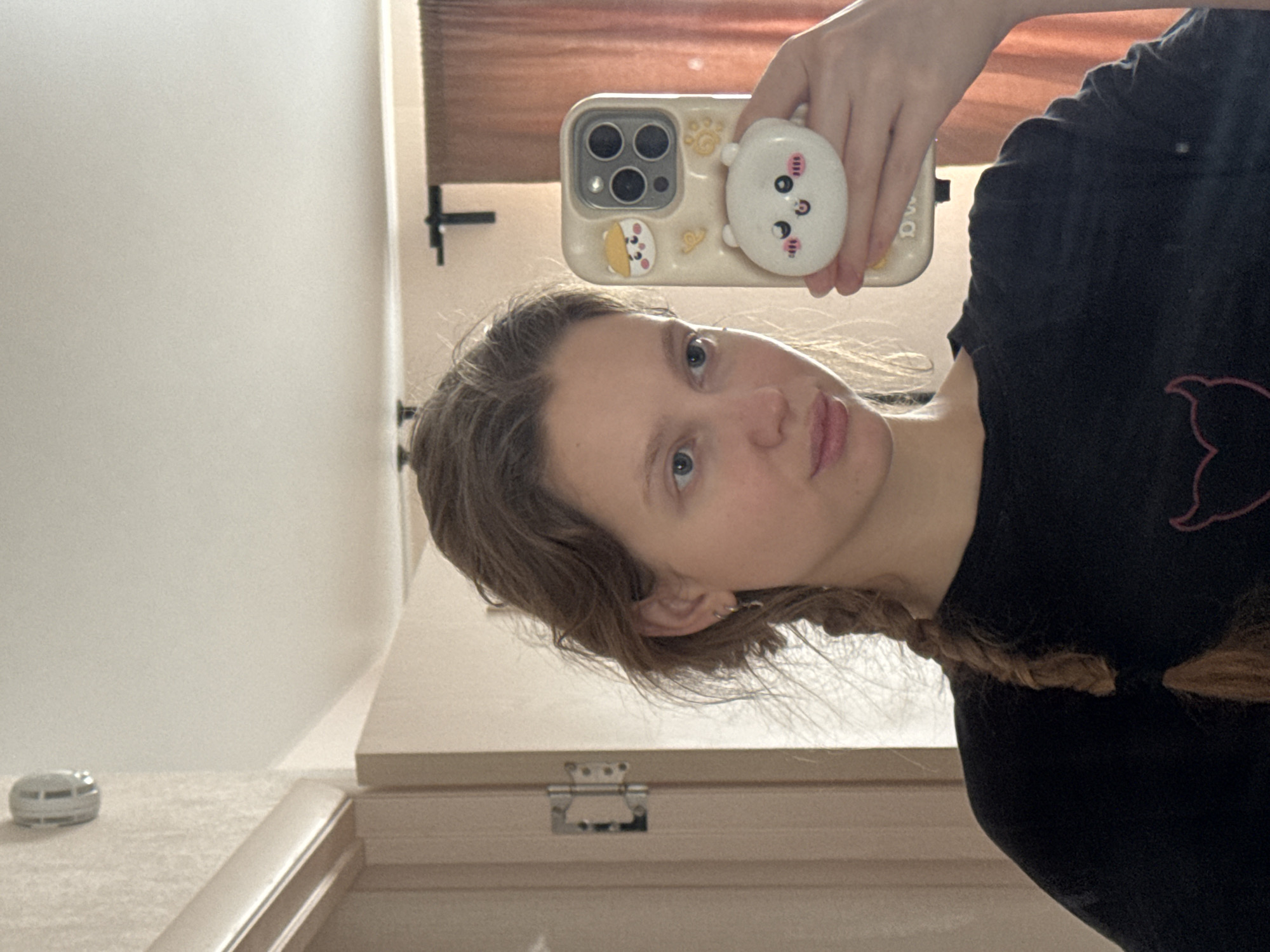 A medium mirror selfie captures a young woman looking directly at the camera, with the image rotated 90 degrees clockwise. The woman has light brown hair, partially styled in a braid over her left shoulder, and is wearing a black t-shirt with a faint pink outline design on the chest. Her expression is neutral.
She holds a cream-colored smartphone (likely an iPhone 14 Pro or Pro Max, identifiable by its triple camera array) encased in a decorated phone cover. The case features small cartoon animal designs, including a yellow character and a larger white character serving as a pop socket, which she is holding. Her fingers are visible wrapped around the phone.
The reflection reveals an indoor setting, possibly a bedroom or dressing area, with warm, indirect lighting suggesting daytime. Visible elements in the background include a light-colored wall, a wooden surface that appears to be a dresser or shelf, a metal hinge from what might be a cabinet door, and a brown-toned reflection that could be blinds or curtains. A small, silver, circular object, possibly a sensor or a small device, rests on the surface to the left. No specific visual cues confirm the provided location context of Izhevsk, Russia, beyond a typical domestic interior. There is no discernible text visible in the image.