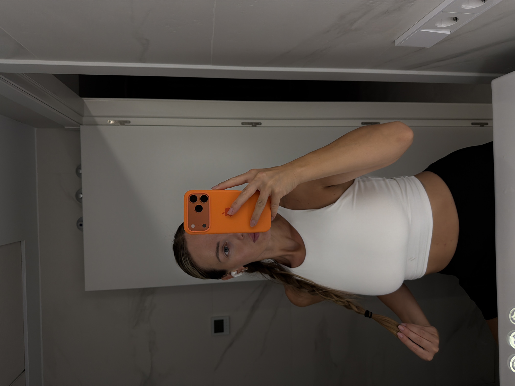 A woman is taking a mirror selfie in what appears to be a bathroom. She is wearing a white crop top and black shorts, with her blonde hair in a braid. She is holding an orange phone up to take the picture. The bathroom has marble-patterned walls and a white door. There are electrical outlets on the wall to the right of the frame. The location is identified as Finestrat, Spain, but this is not visually verifiable from the image itself.