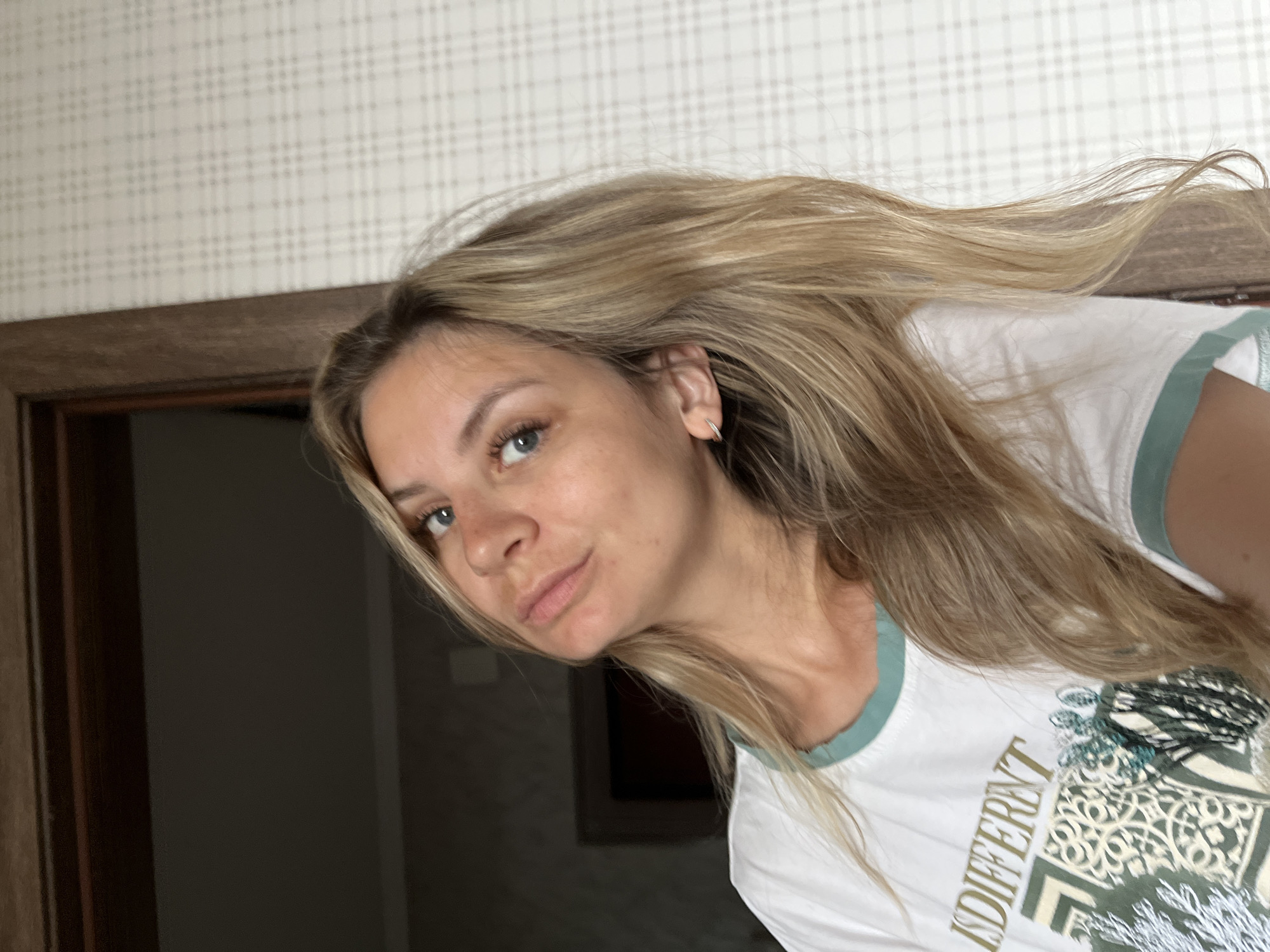 A medium shot captures a Caucasian woman with blonde, wavy hair and blue eyes, looking directly at the camera. She is wearing a white t-shirt with a green neckline and a graphic print. Her head is tilted slightly to her right, and her mouth is slightly parted. The background shows a door frame on the left and a patterned wallpaper on the right. The lighting suggests an indoor environment, possibly a residential room. The location is not definitively identifiable as Pionersky, Russia, based on the provided image alone.