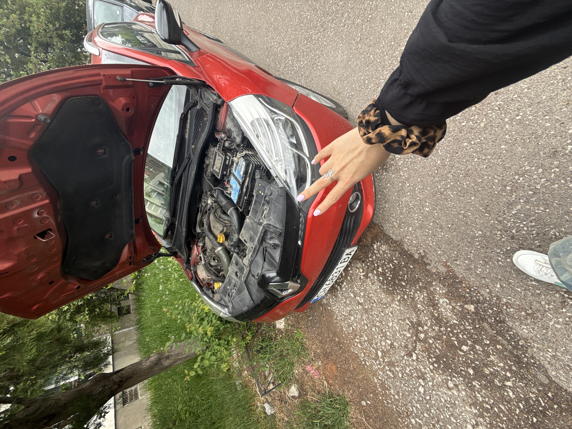A red car with its hood open is parked on a paved surface, likely a road or parking lot, in what appears to be a city setting. The license plate is partially visible and reads "CH 6831 BA." A person is standing near the front of the car, with their hand extended towards the headlight. The person is wearing a long-sleeved black shirt and a scrunchie with a leopard print on their wrist. A silver ring is visible on their finger. The car's engine compartment is exposed, revealing various mechanical components. In the background, there is greenery and a building, suggesting an urban environment. The location is likely in Sofia, Bulgaria, based on the license plate country code.