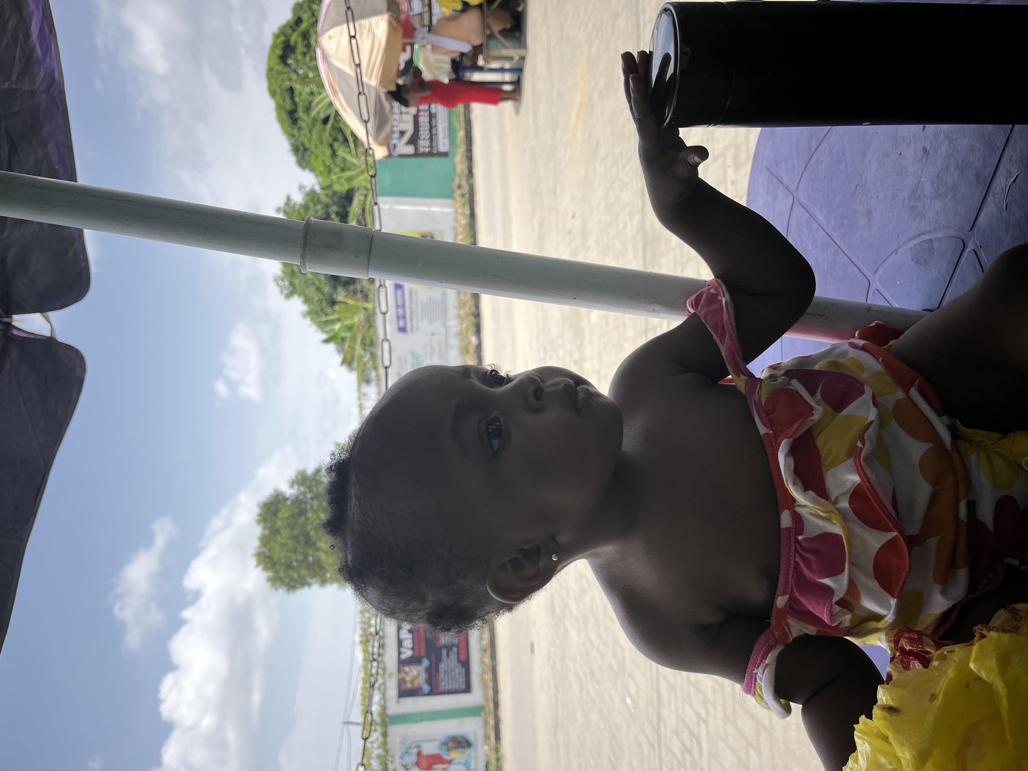 A baby sits on a purple plastic chair outside in Lekki, Nigeria, under a tent structure. The baby wears a colorful floral patterned dress with pink trim and has a small earring. The baby is looking up and touching a black cylindrical object. A white pole runs diagonally across the frame from the upper left to the mid-right. In the background, there are trees, a blue sky with white clouds, and a building with signs. Other people can be seen standing under the tent. The ground is light gray concrete.