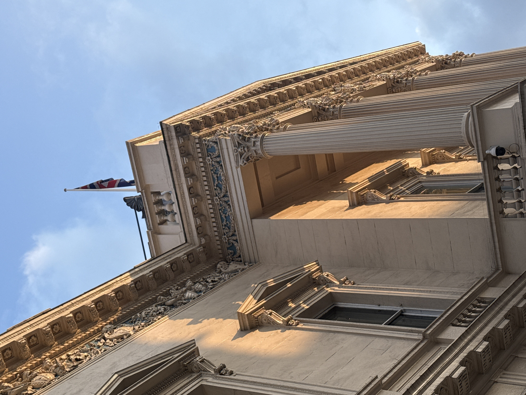 The image is a low-angle view of a richly ornamented classical building in London, United Kingdom. The building is constructed of light-colored stone. The architecture is ornate, with numerous columns, decorative carvings, and window surrounds. A Union Jack flag is flying atop the building. A security camera is affixed to the wall, next to one of the columns. The sky is blue with some clouds.