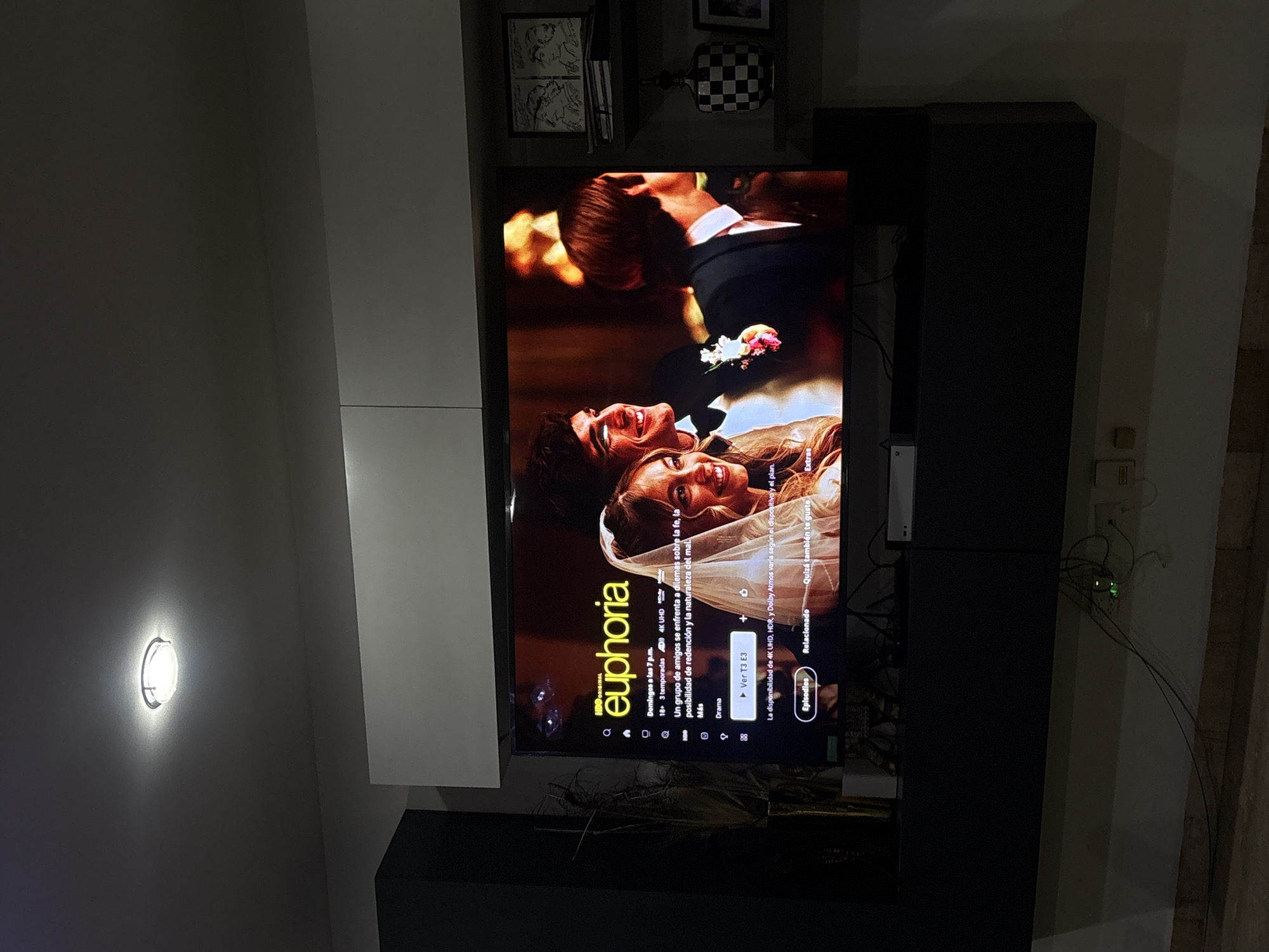 The image shows a television displaying the title screen for the HBO show "Euphoria." The screen features a man and woman smiling and looking at each other, dressed in what appears to be wedding attire. The television is mounted on a wall unit that is partially white and black. Various items are on shelves above the television. The setting appears to be indoors. The precise location of Santa Clara Ocoyucan, Mexico, is not discernible in the image.
