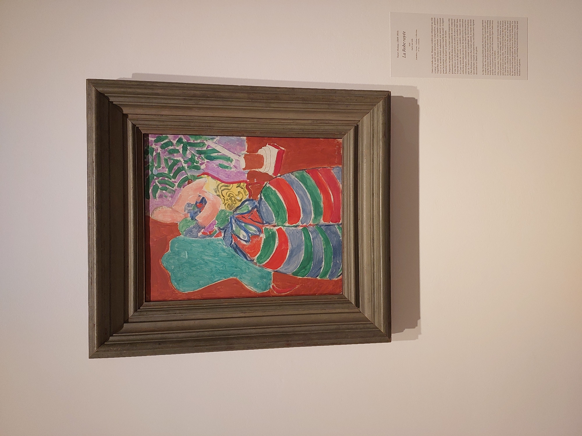 The image shows a framed painting titled "La Robe rayée" by Henri Matisse (1868-1954). The painting itself is a Fauvist work featuring bold colors and simplified forms. It depicts a woman wearing a brightly striped dress, seated in a chair. A plant with purple and green leaves is visible in the background, along with a red book on a table. The painting is displayed on a white wall in what appears to be an art gallery or museum.
Below the painting, a small plaque provides information about the artwork, including its title, artist, date, and collection. The plaque also includes a textual description of the painting in both French and English. The English description states: "In 'La Robe rayée' (1937), Matisse captures an element at the centre: the striped dress, its composition, guiding hand, and rhythm. This garment, almost a second skin, is modelled and rendered with striking visual interest for the public and its painter too. In 1950, Yves Saint Laurent discovered the dress."
While the image depicts a Matisse painting, there is no information within the image itself to indicate its specific location in the city of Nice, France, nor to confirm if the current display is in Nice. The information on the plaque does not provide this geographical context.