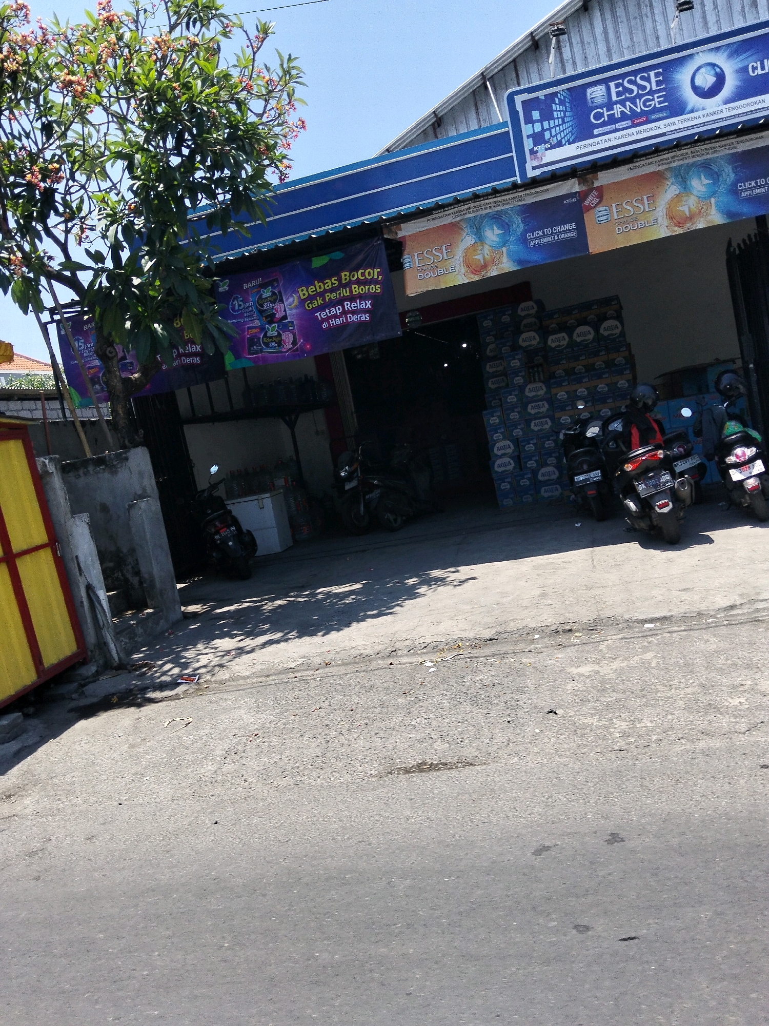In Denpasar, Indonesia, a street-level shot captures a storefront area. In the foreground, a grey asphalt road is visible, with shadows from overhead trees dappling the surface. A weathered, grey concrete wall stands to the left, adjacent to a yellow gate with red trim. Beyond the wall, a street-facing building is adorned with large banners advertising "ESSE CHANGE" cigarettes, featuring images of the product and colorful graphics.
Underneath the banners, the entrance to a shop is partially obscured by a leafy green tree. Inside the shop, stacks of bottled water cases labeled "AQUA" are prominently displayed. Several motorcycles are parked in front of the shop, including a black scooter with license plate "DK 5445 PR" and another black motorcycle with a rider wearing a helmet and a red jacket. Another motorcycle, partially visible, is parked to the left. The overall scene depicts a commercial area on a bright, sunny day.