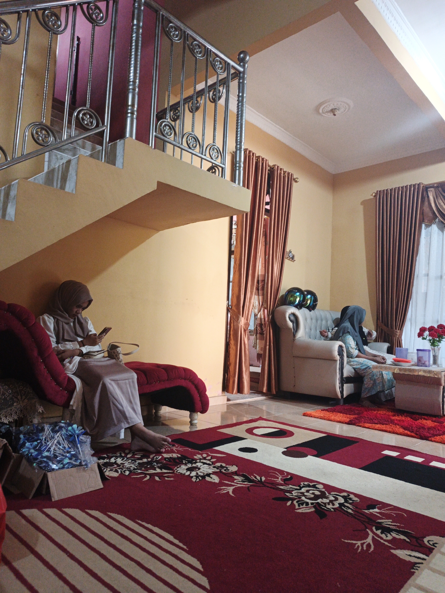 The image shows an indoor scene, likely a living room or common area, in what appears to be a residential dwelling in Pematang Duku, Indonesia. The room is decorated with warm, earthy tones, featuring yellow walls and dark red curtains.
In the foreground, a plush red chaise lounge is occupied by a person wearing a light brown hijab and a long, light-colored dress. They are seated and looking at a mobile phone they are holding in their hands. Beside the chaise lounge, a cardboard box is filled with blue decorative items, possibly flowers or ornaments, wrapped in plastic. A patterned rug with red, black, white, and beige geometric designs covers a significant portion of the floor.
Further into the room, a cream-colored sofa is positioned, with two individuals seated. The person on the left is wearing a blue and white patterned dress and a dark grey hijab. They appear to be looking towards the right. Two green and blue motorcycle helmets are placed on the back of the sofa. To the right of the sofa, a small coffee table holds a vase of red flowers and two purple decorative items. Another rug, with a gradient of red and orange, is placed in front of the coffee table.
A staircase with yellow steps and a silver railing is visible on the left side of the image, leading upwards. The railing features decorative circular patterns. The ceiling is white with subtle decorative molding and a central medallion. The overall impression is of a comfortable, lived-in domestic space.