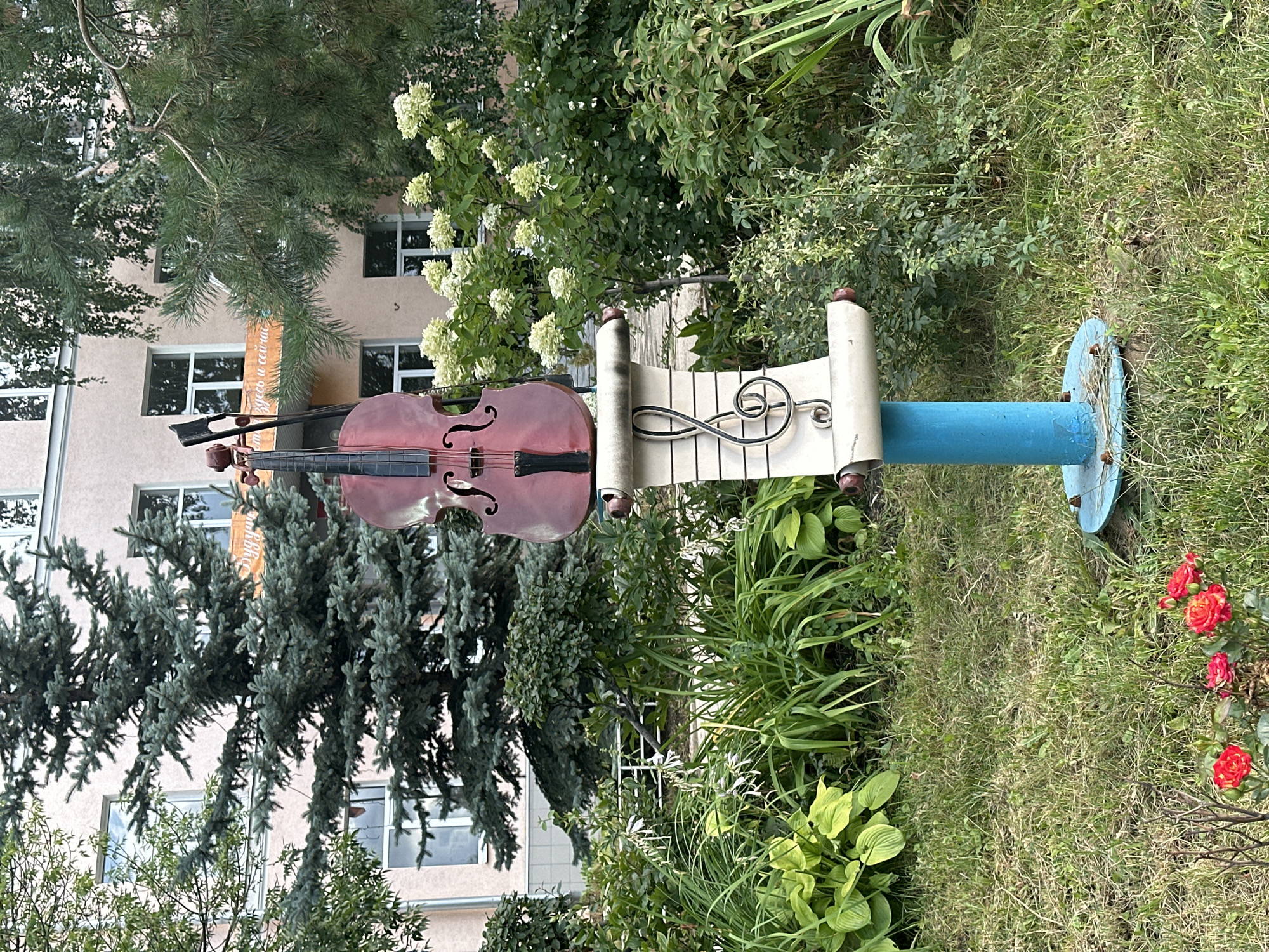 In an outdoor setting with lush green vegetation and a building in the background, a cello and bow are displayed vertically on a blue post topped with a scroll-shaped object adorned with a treble clef. The cello is a reddish-brown color, and the scroll appears to be made of a light-colored material with dark red accents at its ends. The blue post has a circular base. The background features a building with windows and a banner with text in Cyrillic script, as well as various trees and flowering bushes. There are also several red roses in the foreground. The scene appears to be a public space, possibly a park or garden.
