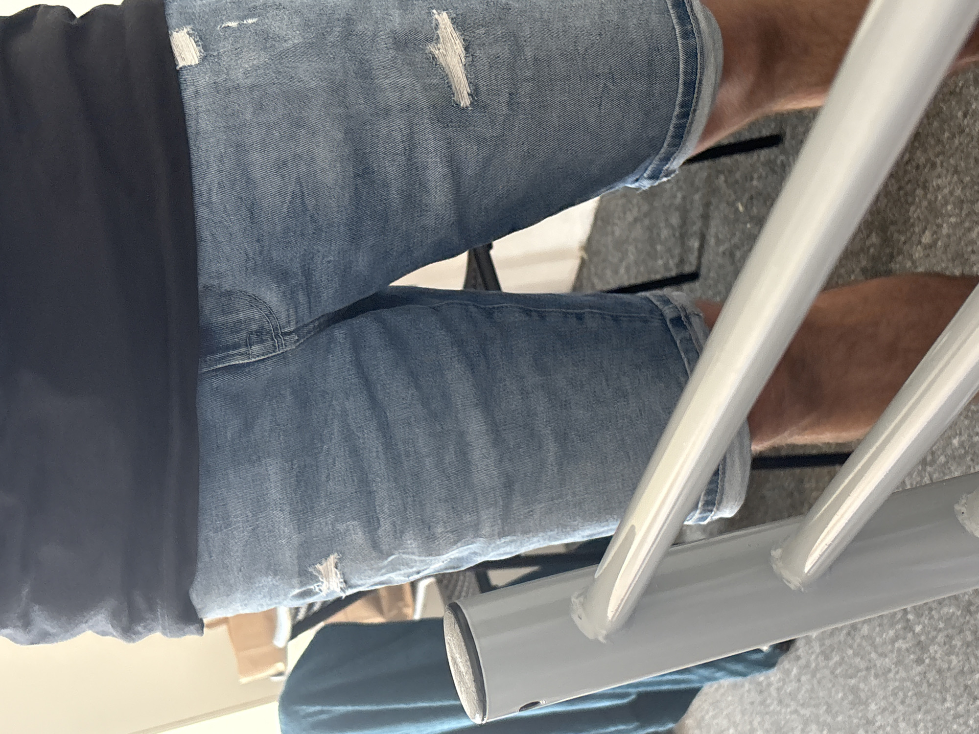 The image shows a close-up of a person's torso and legs wearing a black t-shirt and distressed denim shorts. The shorts are light blue with rips and frayed edges. The person is standing, and their legs are visible from mid-thigh to just above the knee. A light gray metal railing is in the foreground, partially obscuring the lower part of the image. The background is out of focus, but appears to be an indoor setting with some furniture and a blue cloth. The image is taken from a slightly low angle, looking up towards the person's waist. There is no indication of the city or specific location.