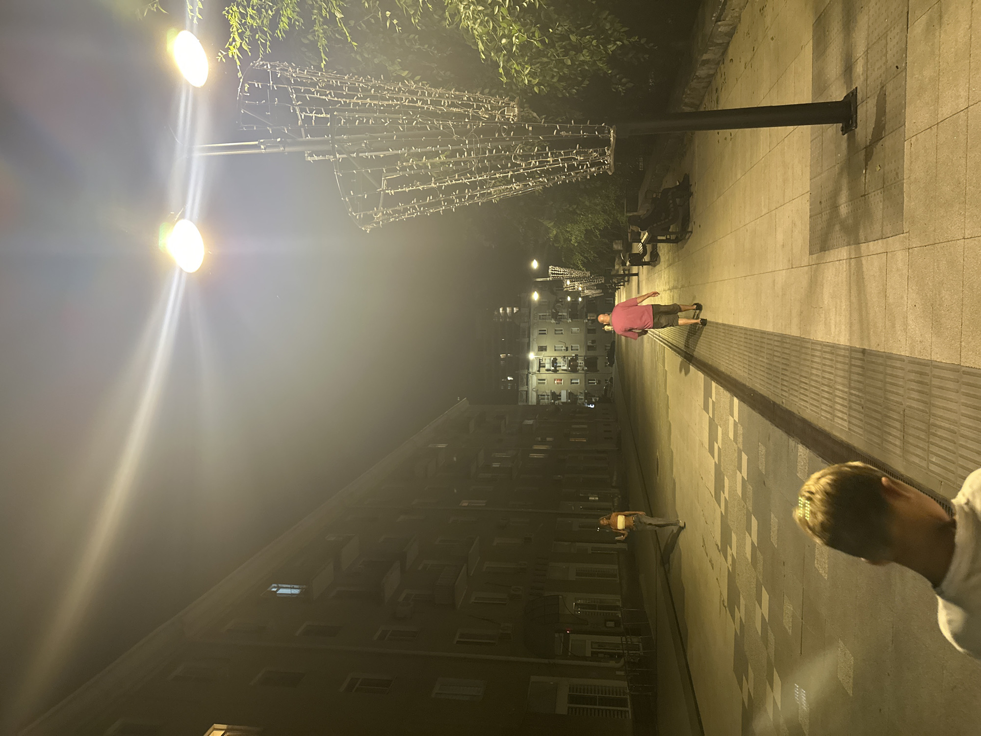 The image depicts a street scene at night in Saratov, Russia, characterized by low visibility due to fog or mist. The street is lined with multi-story buildings on the left and trees and streetlights on the right. A young male is in the foreground, facing away from the camera, with his back to the viewer. Further down the sidewalk, a man in a pink shirt and shorts walks away from the camera. A young woman in a white top and jeans walks towards the camera in the distance on the left side of the street. The sidewalk is paved with tiles, and there are benches along the right side of the path. Decorative light fixtures are mounted on tall poles, illuminated by bright streetlights.