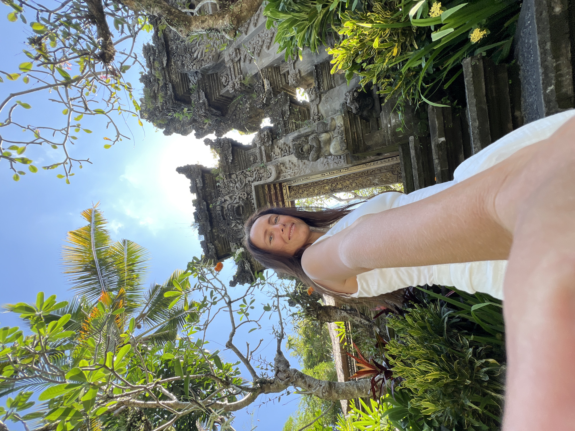 A selfie of a Caucasian woman with long brown hair, wearing a white top, is taken from a low angle in Kedewatan, Indonesia. The background features an ornate, stone Balinese gateway structure overgrown with moss and vegetation. Lush greenery, including trees with broad leaves, and a palm tree with yellow fronds, frame the blue sky. The sun is visible above the gateway, creating a bright, diffused light.