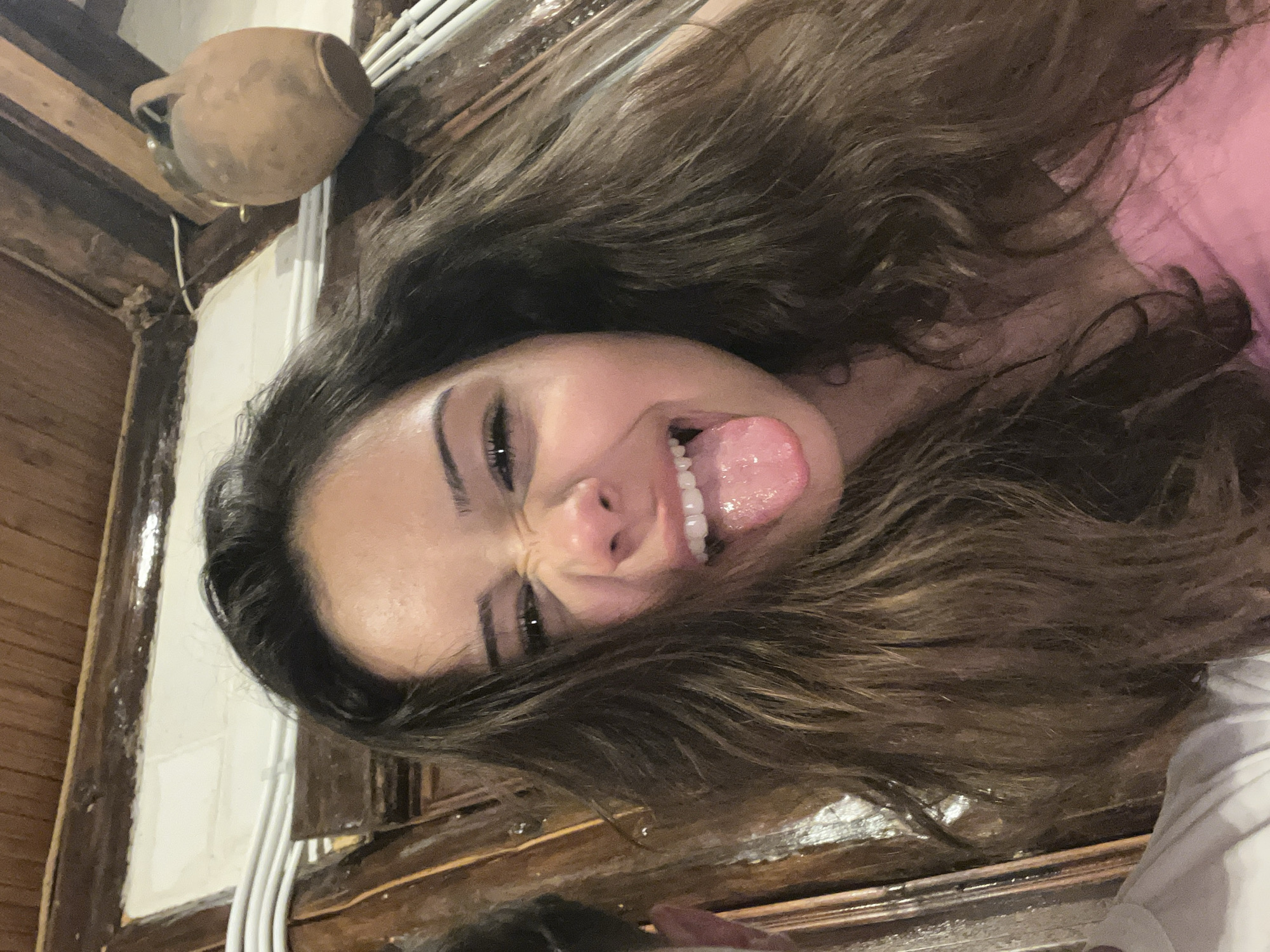 A woman with long, brown hair is indoors, sticking her tongue out and making a face at the camera. The background appears to be a rustic interior with wood paneling, white pipes, and a clay pot hanging on the wall. The location is Presoka, Bulgaria.