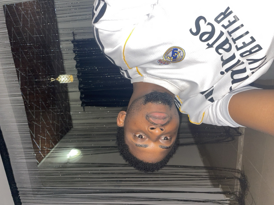 A medium close-up, tilted photograph captures a single male individual of African descent, appearing approximately 20-30 years old, looking directly at the camera with a neutral expression. He has short, dark, curly hair and a short, neatly trimmed dark beard. He is wearing a white short-sleeved Real Madrid jersey with black Adidas stripes on the shoulders and yellow trim. The Real Madrid club crest is visible on the left chest, and the sponsor "Emirates FLY BETTER" is printed in black across the front.
The subject is positioned indoors against a dimly lit background. Above and behind him, a dark ceiling with a reflective, geometric patterned panel is visible, from which a decorative, illuminated gold-toned chandelier hangs. Dark, translucent string or bead curtains are present vertically across the background, partially obscuring what appears to be a darker fabric curtain on the right. A section of a light-colored floor is visible at the bottom left. The image provides no discernible unique geographic markers to confirm the location within Ube, Nigeria; the setting is an unidentified interior space.
