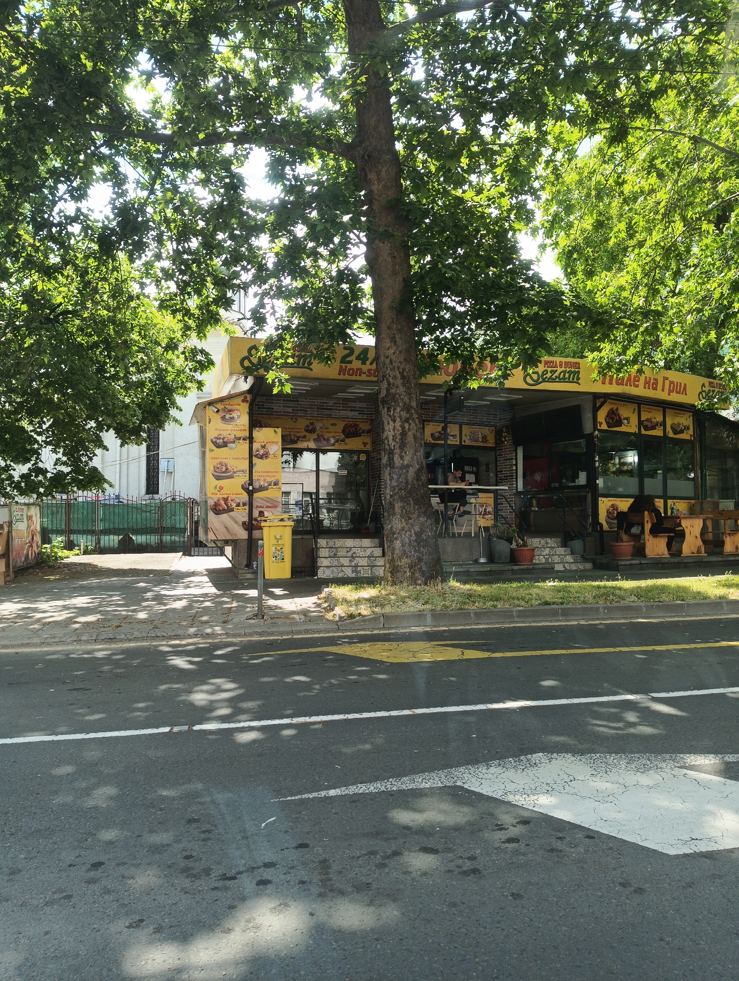 Here is a detailed description of the image:
**Scene Overview:** The image shows a street scene in what appears to be Burgas, Bulgaria, based on the Cyrillic text visible on the storefront. The focus is on a food stand/restaurant named "Sezam" that offers pizza and grill items. The stand is located along a street with a crosswalk in the foreground.
**Environment:** The scene is partially shaded by trees, with sunlight filtering through the leaves creating dappled patterns on the road and sidewalk. There is a building behind the food stand on the left, with a green fence and gate.
**Food Stand/Restaurant Details:**
* The stand has a yellow awning with menu items displayed in Cyrillic, along with images of food items.
* The restaurant's signage includes "24h Non-Stop," indicating it's open around the clock.
* Menu items include "Pizza & Doner," and "але на Грил" (Grill).
**People and Actions:** There is a person sitting inside the food stand at a table. Another person is seen outside the food stand.
**Road and Street Details:**
* A crosswalk with white arrow markings is visible on the road.
* There is a yellow line painted along the edge of the road near the sidewalk.
* Shadows from the trees are cast across the road surface.
**Objects:** There is a yellow trash can next to the building. Wooden tables and benches are located outside the food stand for customers. A large tree is positioned directly in front of the food stand, partially obscuring it.
**In summary,** the image captures a moment at a 24-hour food stand "Sezam" in Burgas, Bulgaria, showing the establishment during daylight hours with a mix of natural light and shadow, as well as people and various physical details of the setting.