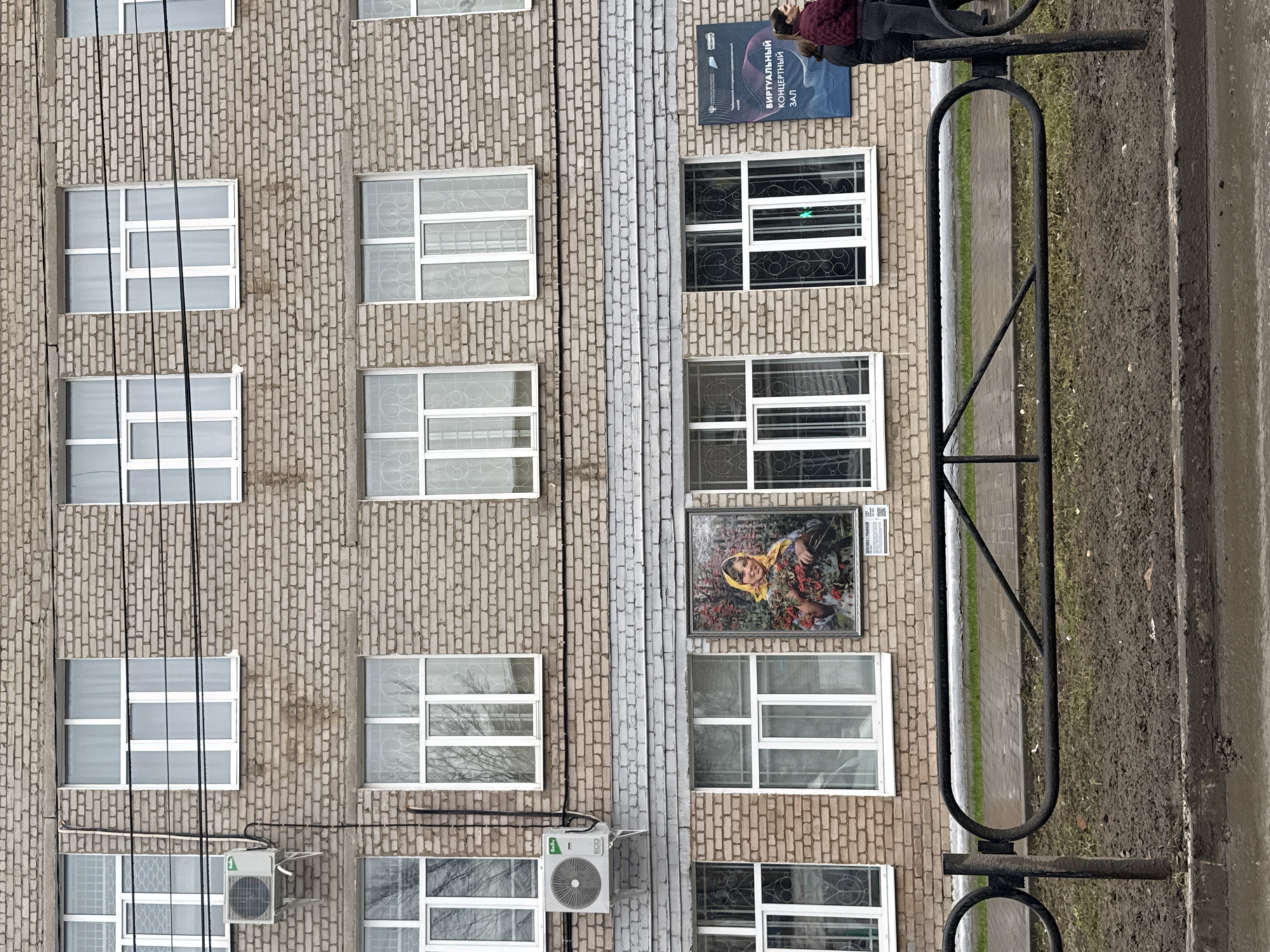 The image shows the exterior of a multi-story brick building. The facade includes multiple windows, and some have decorative metal bars. There is a sign with text in Russian attached to the wall, near the top of the building. An outdoor air conditioning unit is attached to the wall. A large framed painting hangs on the wall between two windows. A metal fence and a sidewalk are visible in the foreground. There is a person near the top of the building, near the sign. The location is Chaikovsky, Russia.