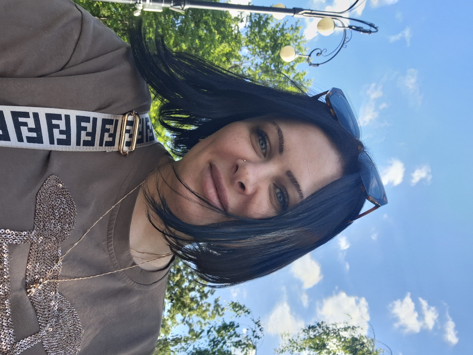 A medium close-up, rotated 90 degrees clockwise, captures a woman taking a selfie outdoors in Moscow, Russia. The woman, with dark hair and green eyes, looks directly at the camera with a gentle smile. She has a small stud piercing on her left nostril. Sunglasses with tortoise-shell frames are perched on top of her head.
She is wearing a short-sleeved, grey-khaki colored top, which features a large, intricate design made of shimmering silver sequins on the chest. A delicate gold chain necklace is visible around her neck. Across her right shoulder (appearing on the left side of the rotated image) is a prominent white strap with the black, repeating "FENDI" logo. A gold-toned buckle on the strap is also visible.
The background shows a bright blue sky with scattered white clouds, suggesting sunny weather during the day. Green foliage from trees is visible, indicating a park or outdoor setting. A black decorative lamp post with spherical lights can be seen against the sky and trees. The overall impression is a casual, pleasant outdoor moment.