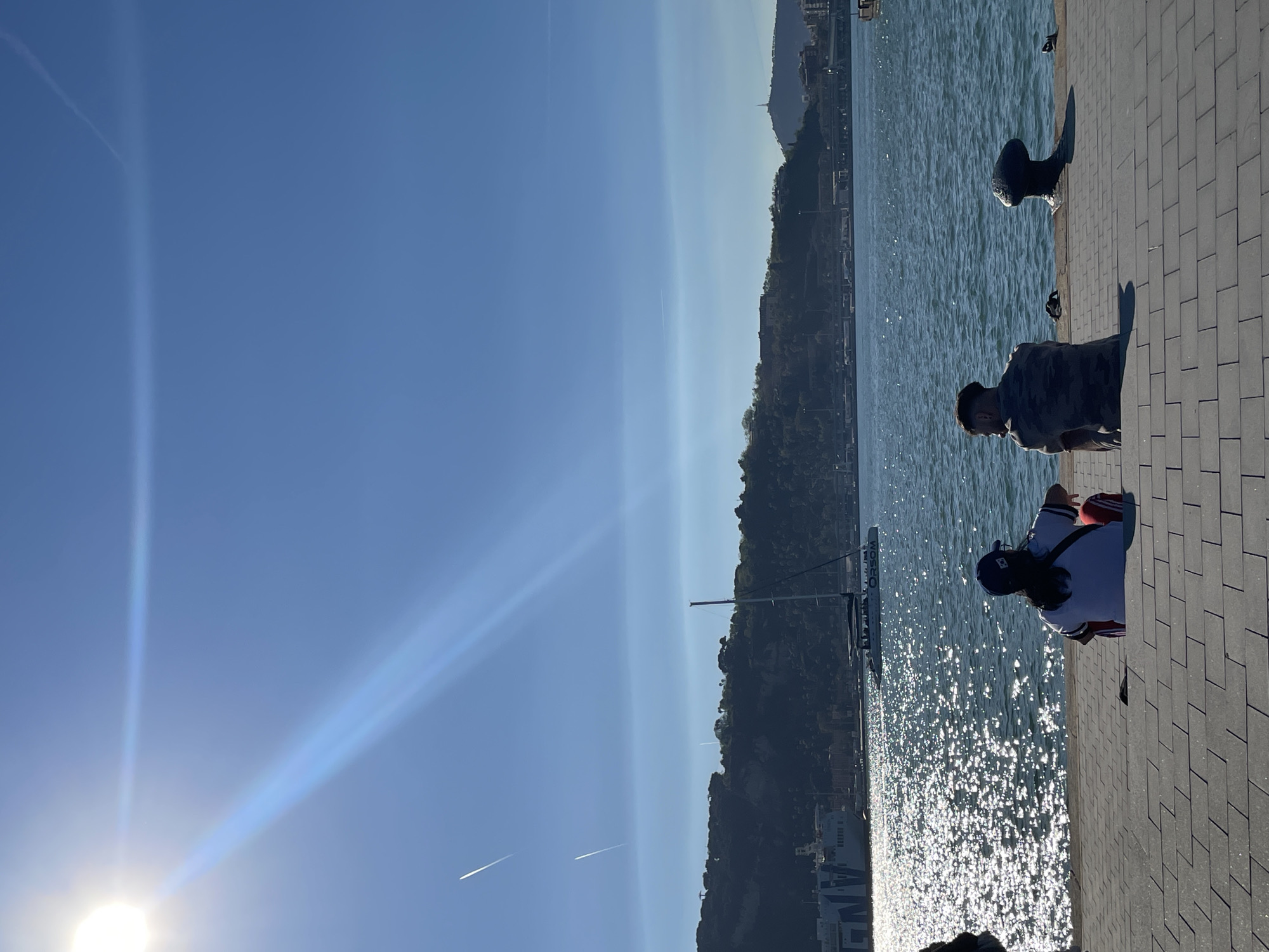 This media file captures a sunny waterfront scene, likely a port or marina in Barcelona, Spain. The sky is a clear, brilliant blue, with the bright sun prominently featured in the upper left, emitting a strong lens flare that streaks across the sky. Two faint airplane contrails are also visible high above.
In the foreground, a paved stone dock or promenade occupies the right side of the image. Adjacent to it, the water surface is intensely bright with myriad sparkling reflections from the sun, indicating a clear, sunny day. Several dark-colored bollards are spaced along the edge of the dock.
Two individuals are on the dock. A man in a camouflage t-shirt stands on the right, facing away from the camera and looking towards the water. Further to the right and lower, another person, with long hair and wearing a dark cap and a white and red shirt or jacket, is seated on the ground, also facing away from the viewer. Both figures are partially silhouetted against the bright water.
In the middle distance, a large body of water extends towards a hilly or mountainous coastline in the background. These hills are covered in vegetation and scattered buildings. A large boat or ship is visible in the water between the foreground and the distant hills. The name "ORSOM" is clearly legible in white letters on its hull, with a partial word "ANG" below it. The bright, high sun suggests it is mid-morning to late afternoon. The scene conveys a relaxed and pleasant atmosphere.