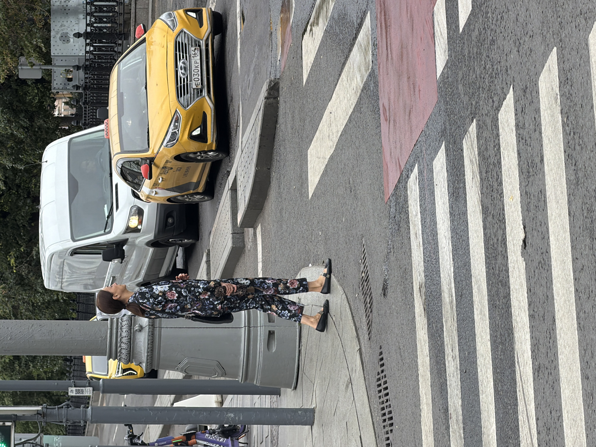 A woman in a floral suit and black flats stands on a crosswalk at a street intersection in Moscow, Russia. She is looking to her right. Behind her, a yellow Hyundai taxi is stopped in traffic next to a white van. The street is wet, suggesting it has recently rained. The crosswalk is marked with white stripes, and a section of red paving is visible at the edge of the crosswalk. A lamppost stands to the left of the woman.