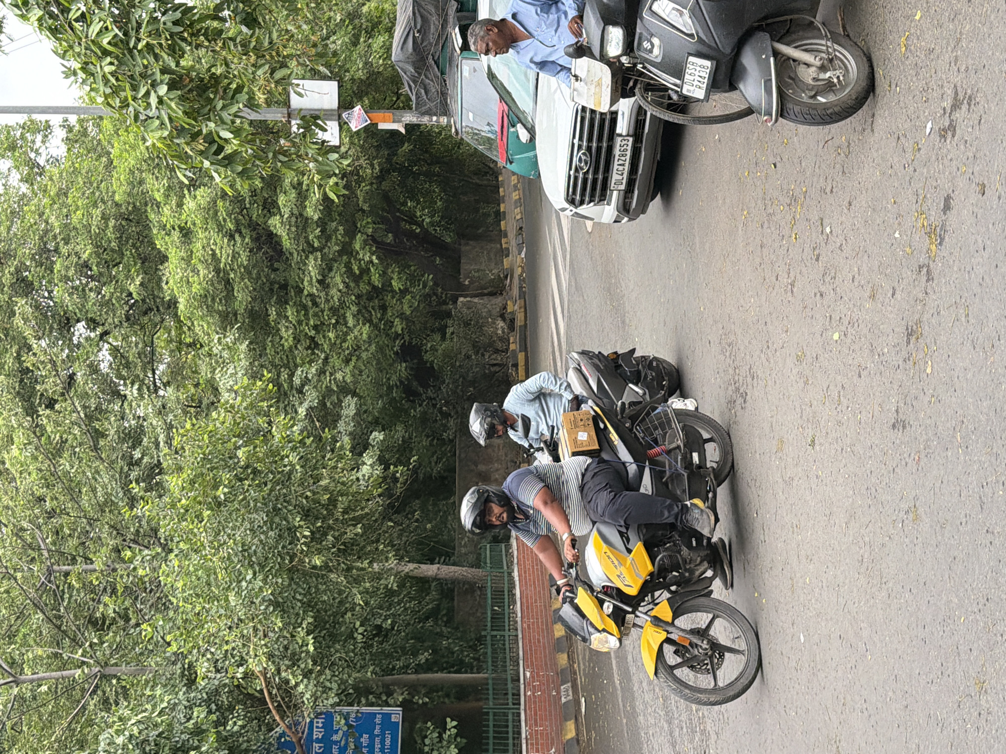 In New Delhi, India, on a street lined with trees and a brick wall, two men ride a yellow motorcycle. The driver, wearing a striped shirt and helmet, steers the bike, while the passenger, in a light blue shirt and helmet, sits behind him holding a cardboard box. In the background, a white car with license plate DL4CAZ8653 is stopped at an intersection. A green truck with a tarp-covered load is behind the car. A black scooter with license plate DL6SB R4438 is also parked nearby, and a man in a blue shirt stands next to it.