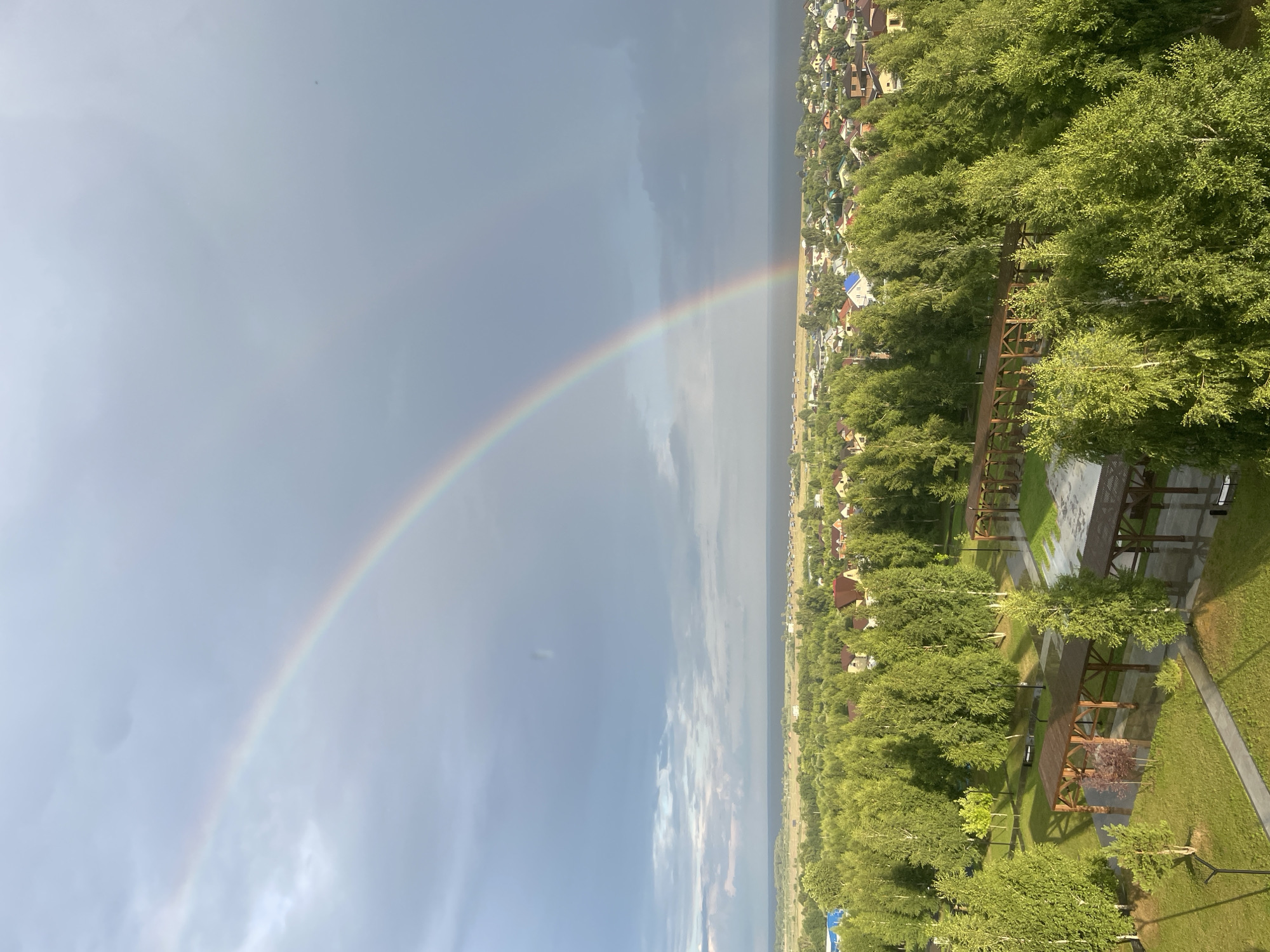 An aerial view captures a double rainbow arcing across a dramatic sky, dominating the upper portion of the image. The sky transitions from a bright, light grey on the right to a darker, stormy grey on the left, with visible cloud formations. Below the sky lies a vast expanse of lush green trees that forms a dense canopy.
Nestled within the treeline, a settlement of residential houses with red and beige roofs is visible, suggesting a semi-rural or suburban setting. Further in the distance, a body of dark water, possibly a lake or sea, separates the settlement from a hazy horizon line.
In the foreground, a park-like area features paved walkways, manicured green lawns, and several wooden structures that appear to be covered pavilions or gazebos. The ground around these structures is wet, indicating recent rainfall. The trees in this foreground area are vibrant green, with some appearing to be birch trees, their white bark visible. The overall scene suggests a post-rain environment in the city of Laishevo, Russia, with the natural beauty of the landscape enhanced by the striking atmospheric phenomenon of the double rainbow. There are no people or animals visible in the image.