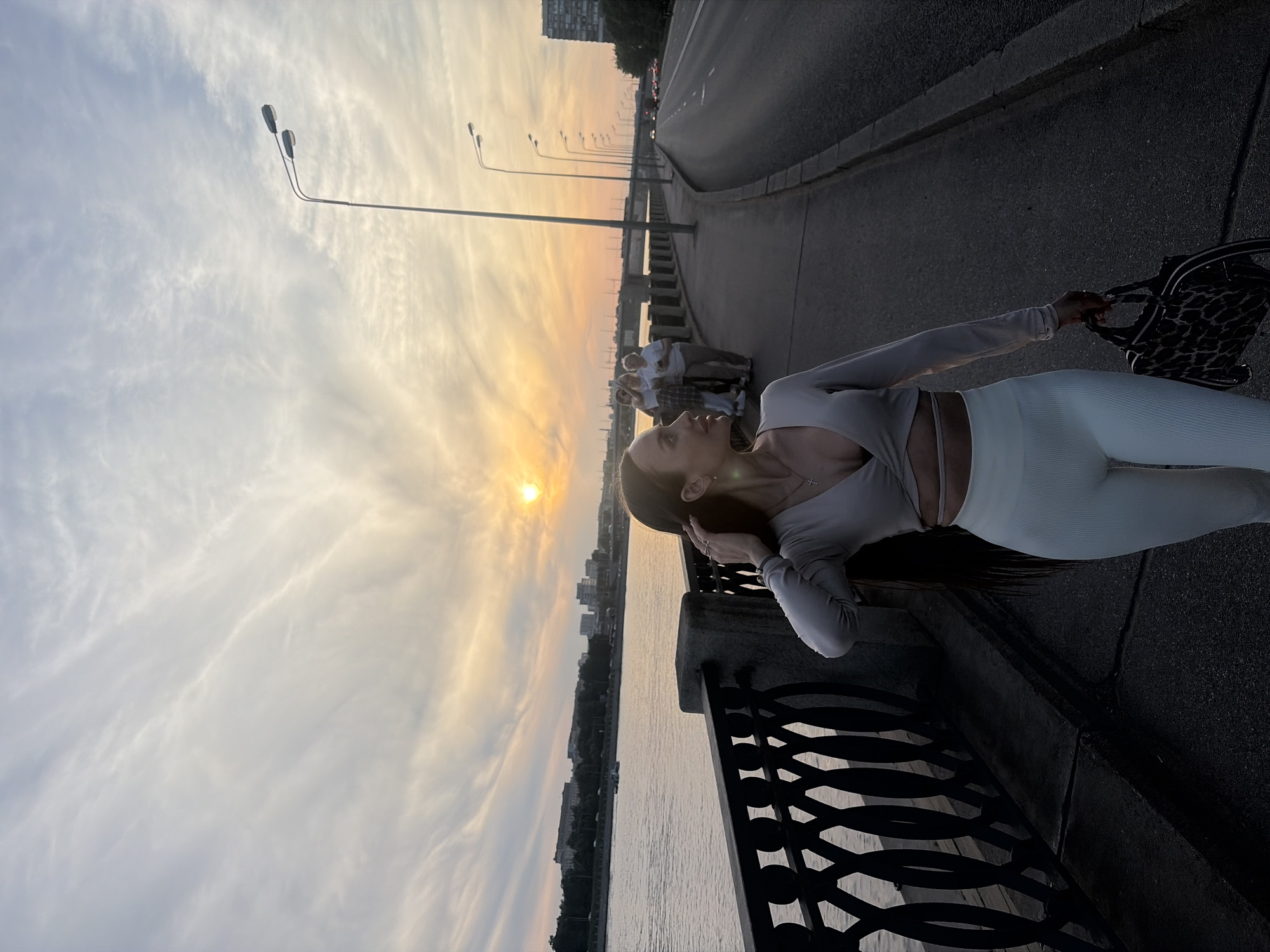 A young adult female is captured walking along a paved riverside promenade during sunset in a city identified as Unknown, Russia. The sun, a bright orange orb, is low in the western sky, casting a warm, golden glow on cirrus clouds and illuminating the scene.
The female subject is positioned in the foreground, walking towards the left side of the frame. She has long dark hair, parted on the side, and is looking slightly to her right, away from the camera. She wears a light-colored, long-sleeved crop top with a V-neck and a wrap-around waist detail, paired with light-colored, ribbed leggings. A dark-patterned handbag, possibly leopard print, is held in her left hand, which is swung slightly forward. Her right hand is raised to her hair near her ear. She wears earrings and a small cross pendant on a necklace.
To the subject's left, a decorative black metal fence with intertwined oval patterns separates the promenade from a wide river. A small boat is visible on the river further in the distance. Across the river, urban buildings and a distant bridge define the skyline, partially obscured by trees.
To the subject's right, a multi-lane asphalt road runs parallel to the promenade. Numerous tall streetlights with dual luminaires line the edge of the road, extending into the background. Vehicles, indicated by faint lights and shapes, are visible on the road in the distance.
Further down the promenade, behind the main subject, a group of three to four individuals are standing and conversing, partially visible between the main subject and the streetlights. The overall lighting is dimming, characteristic of dusk or early evening, with the brightest light emanating from the setting sun.